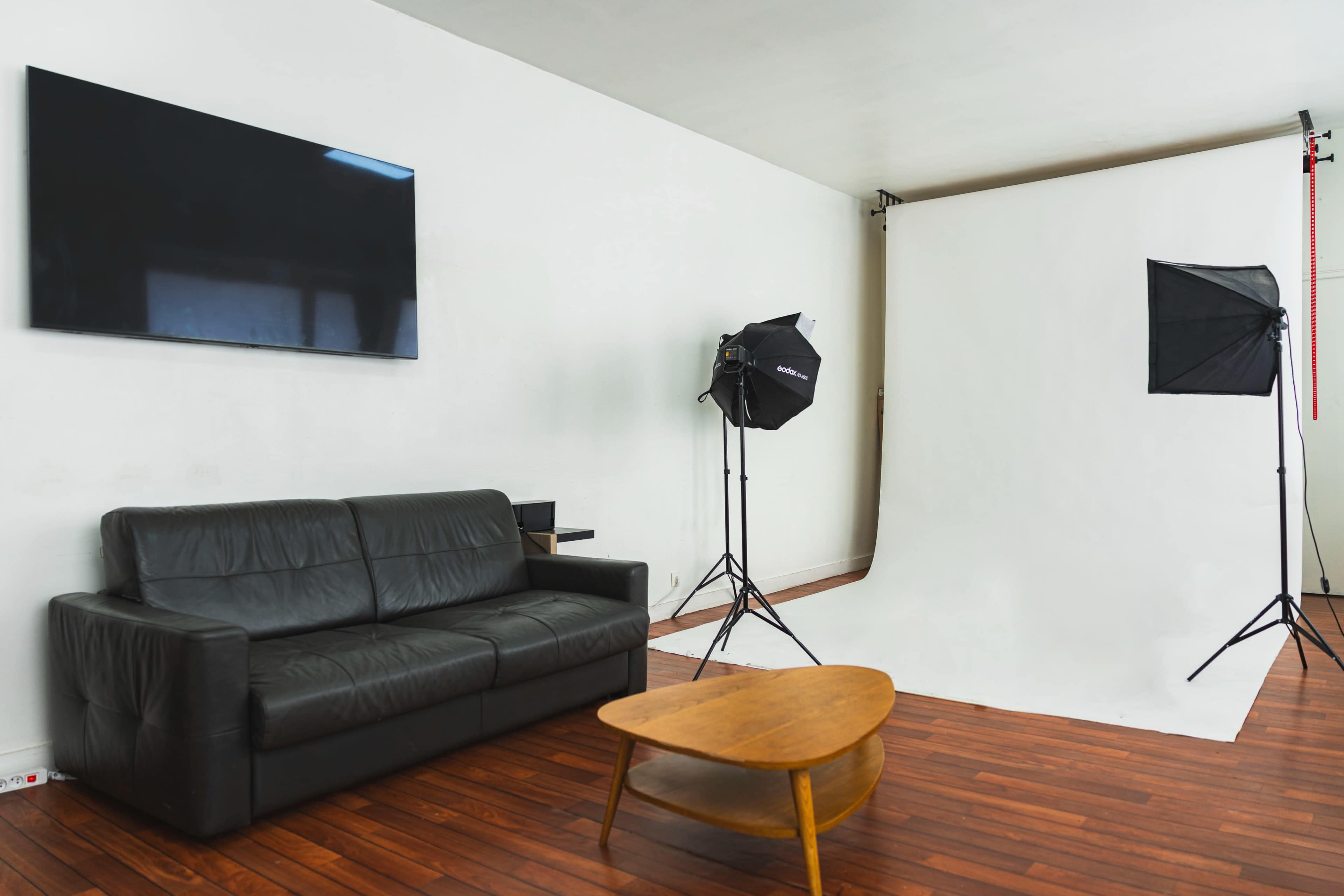 The image shows a bright studio setup with a black leather couch, a wooden coffee table, and two softbox lights near a white backdrop.