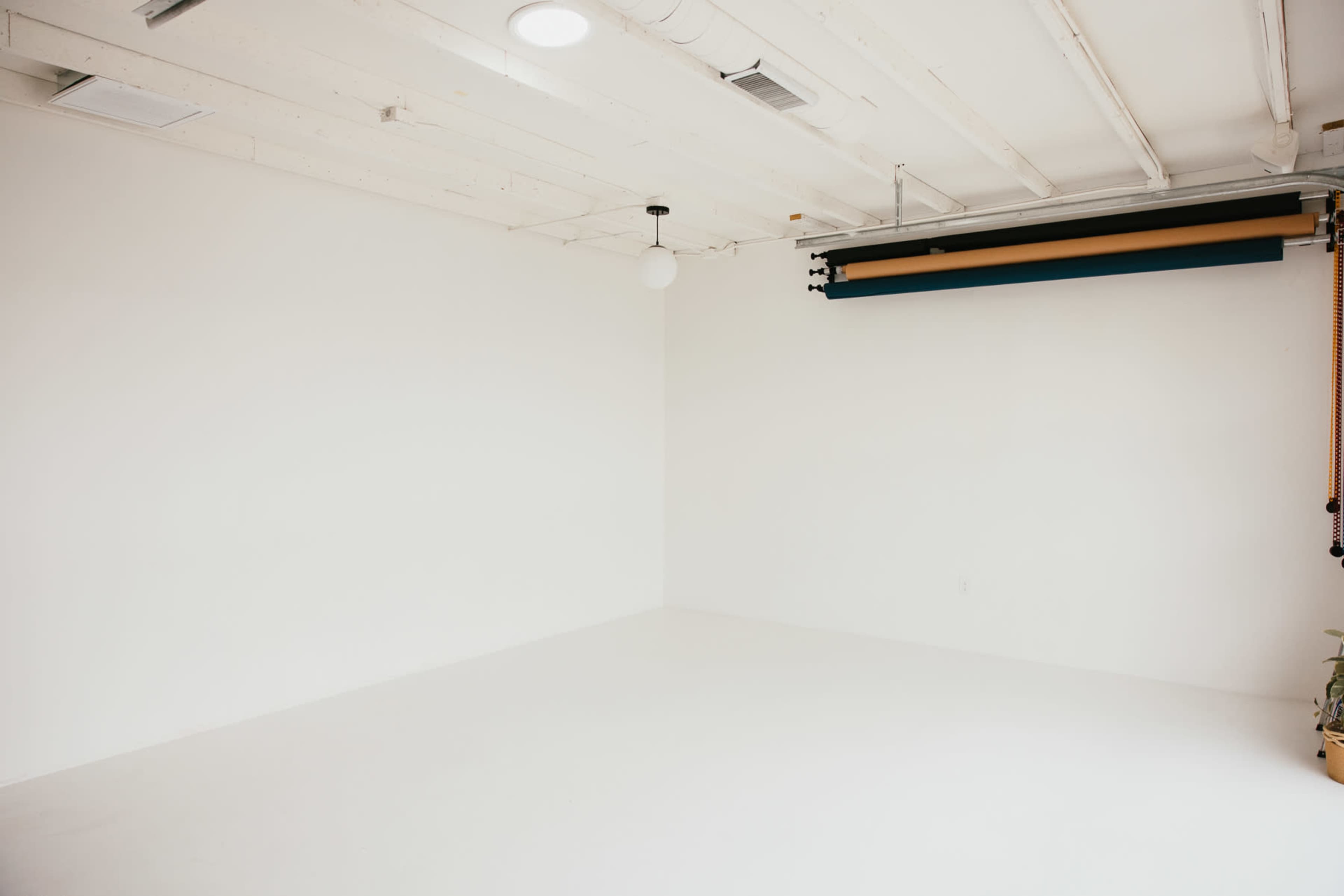 The image shows an empty room with plain white walls and a smooth, light-colored floor, featuring a hanging light fixture and a roll of colored backdrop material on the ceiling.