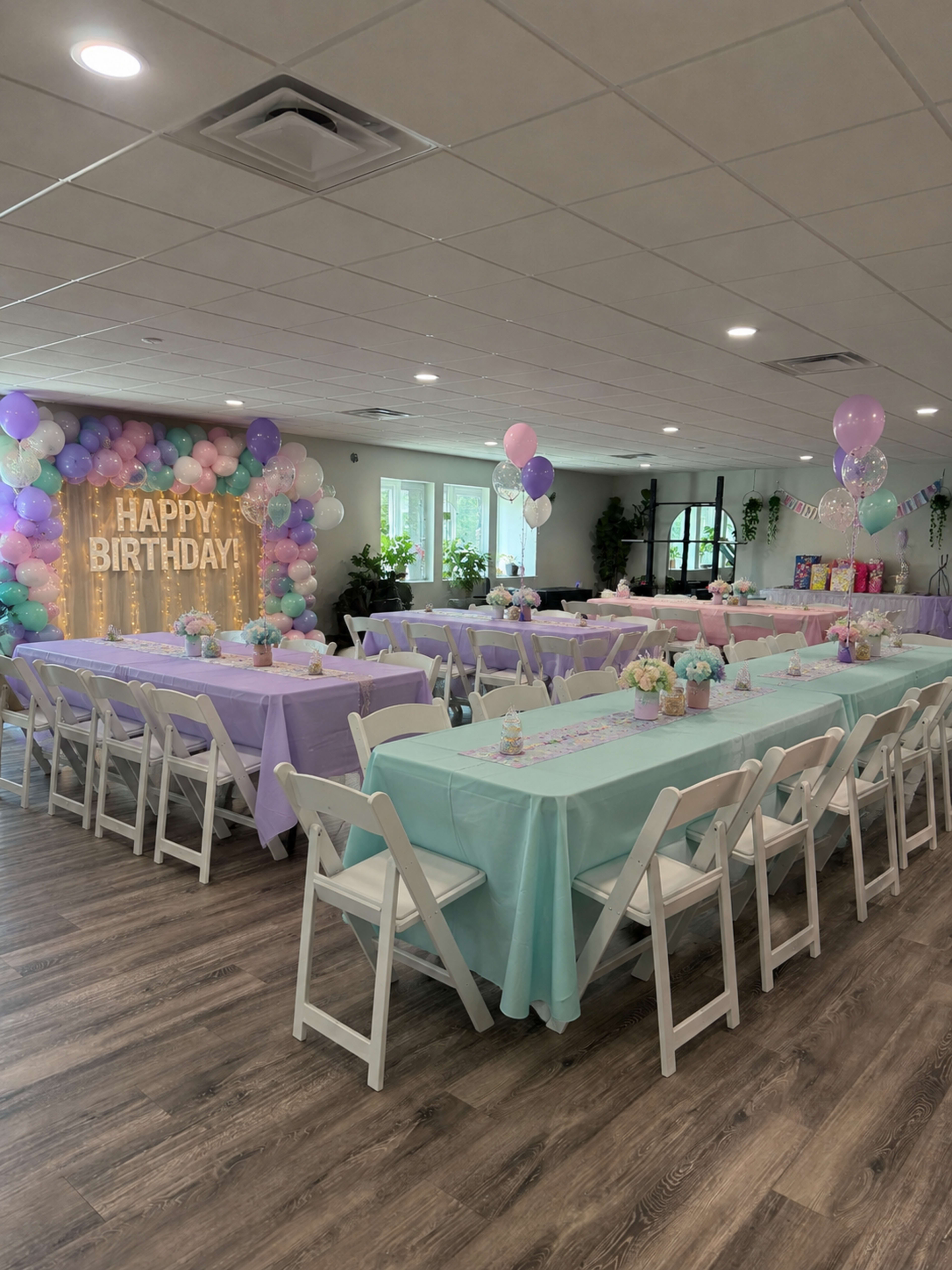 The image shows a decorated party space with tables set in alternating shades of lavender and mint green, surrounded by balloons and a "HAPPY BIRTHDAY" sign on the wall.