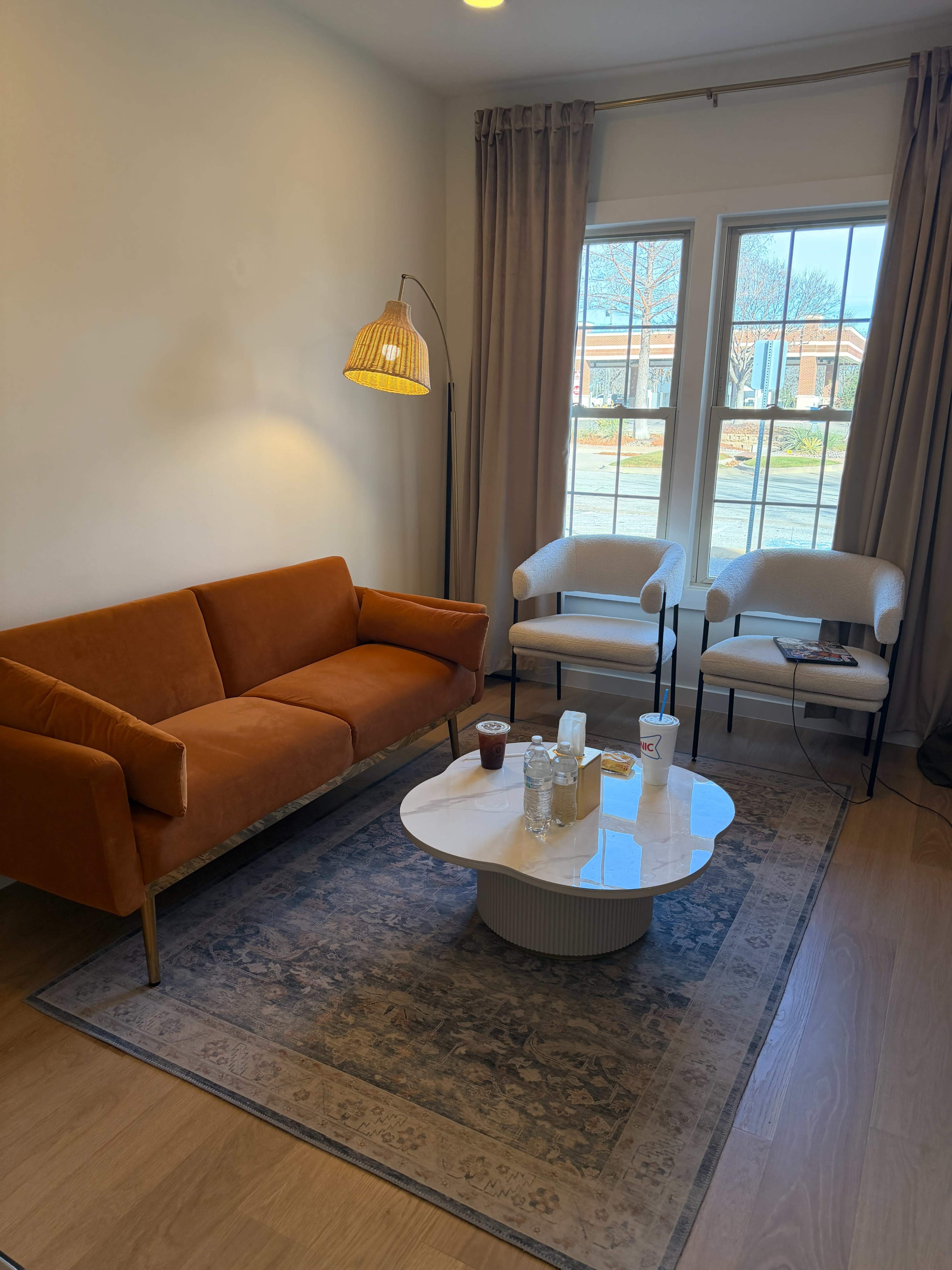 The image shows a cozy living space with an orange sofa, two white chairs, and a round coffee table on a patterned rug beneath a large window.