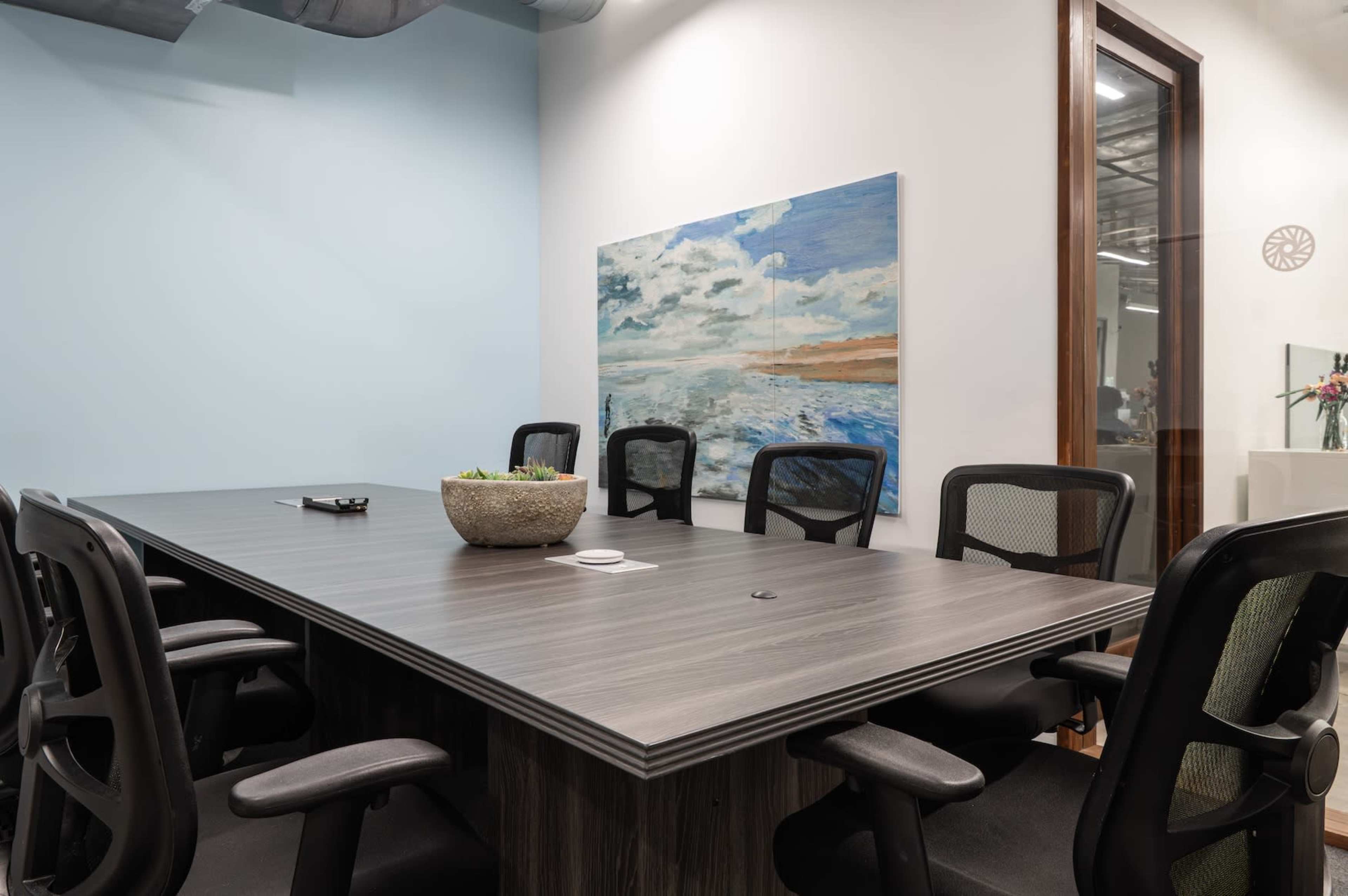 The image shows a conference room featuring a large table surrounded by several black office chairs and a colorful abstract painting on the wall.