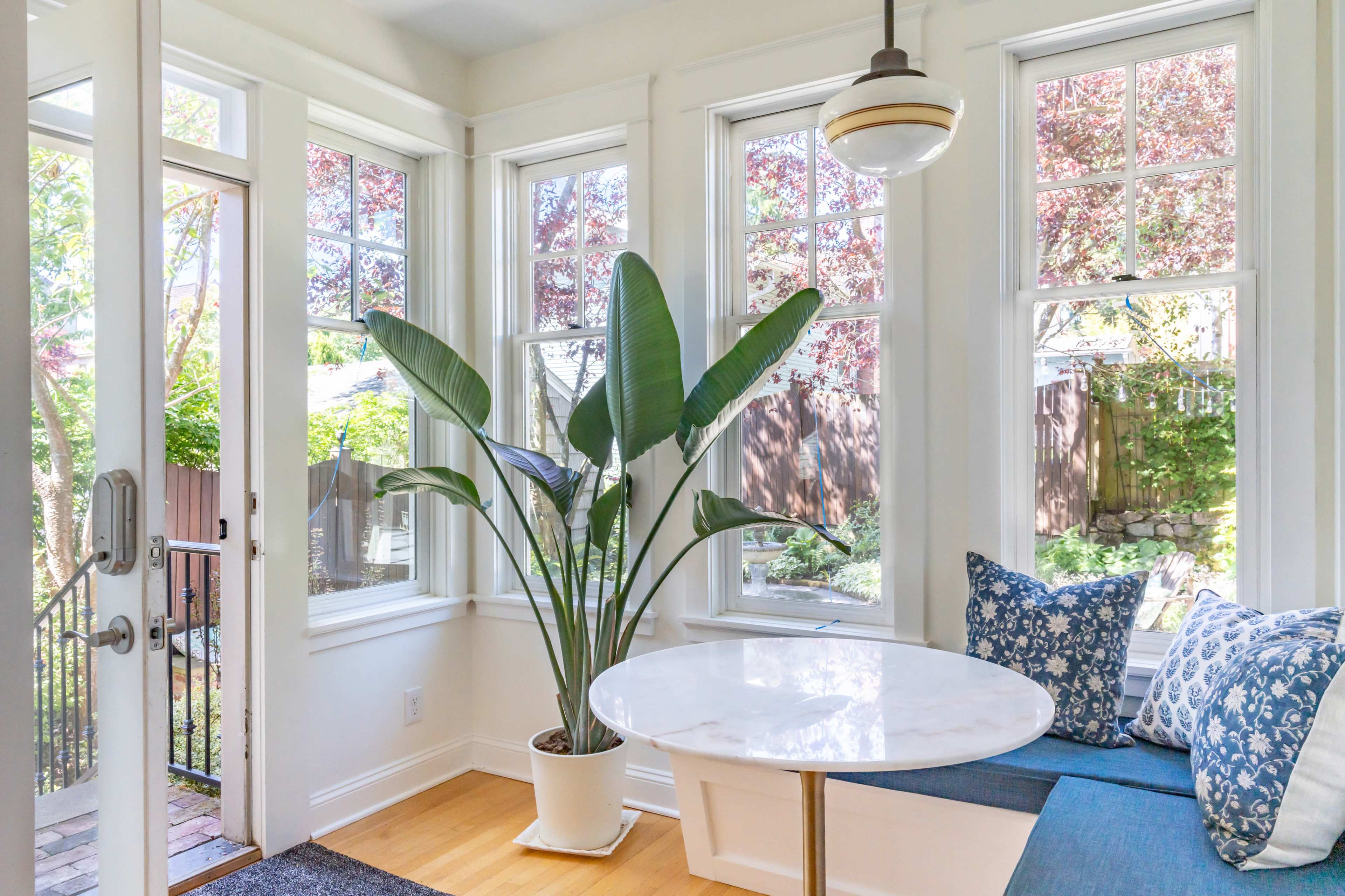 A bright sunroom features a round marble table, a large potted plant, and windows with views of a garden outside.