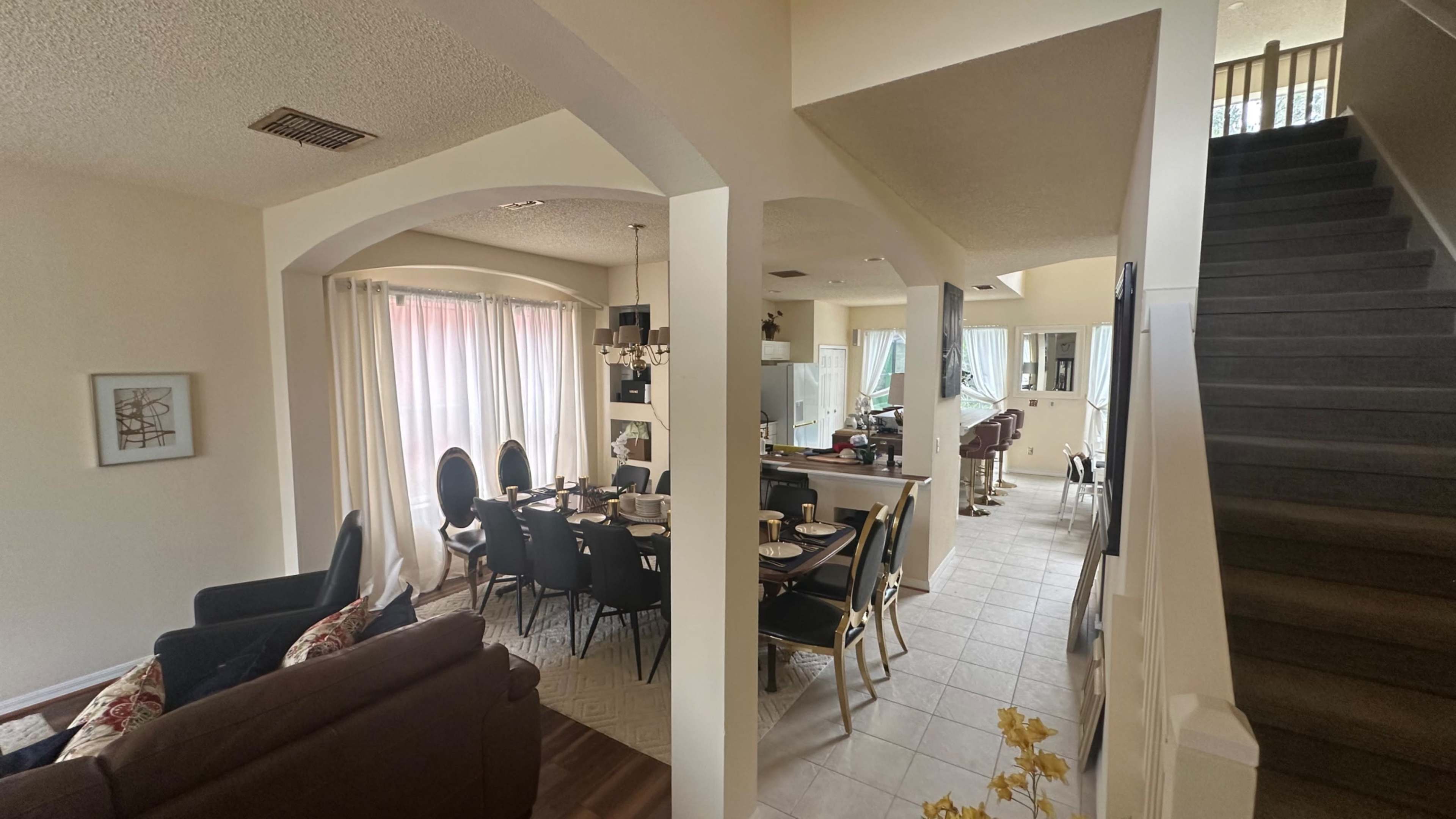 The image shows an open-concept living space with a dining area featuring a black table and chairs on one side and a staircase leading to an upper level on the other.