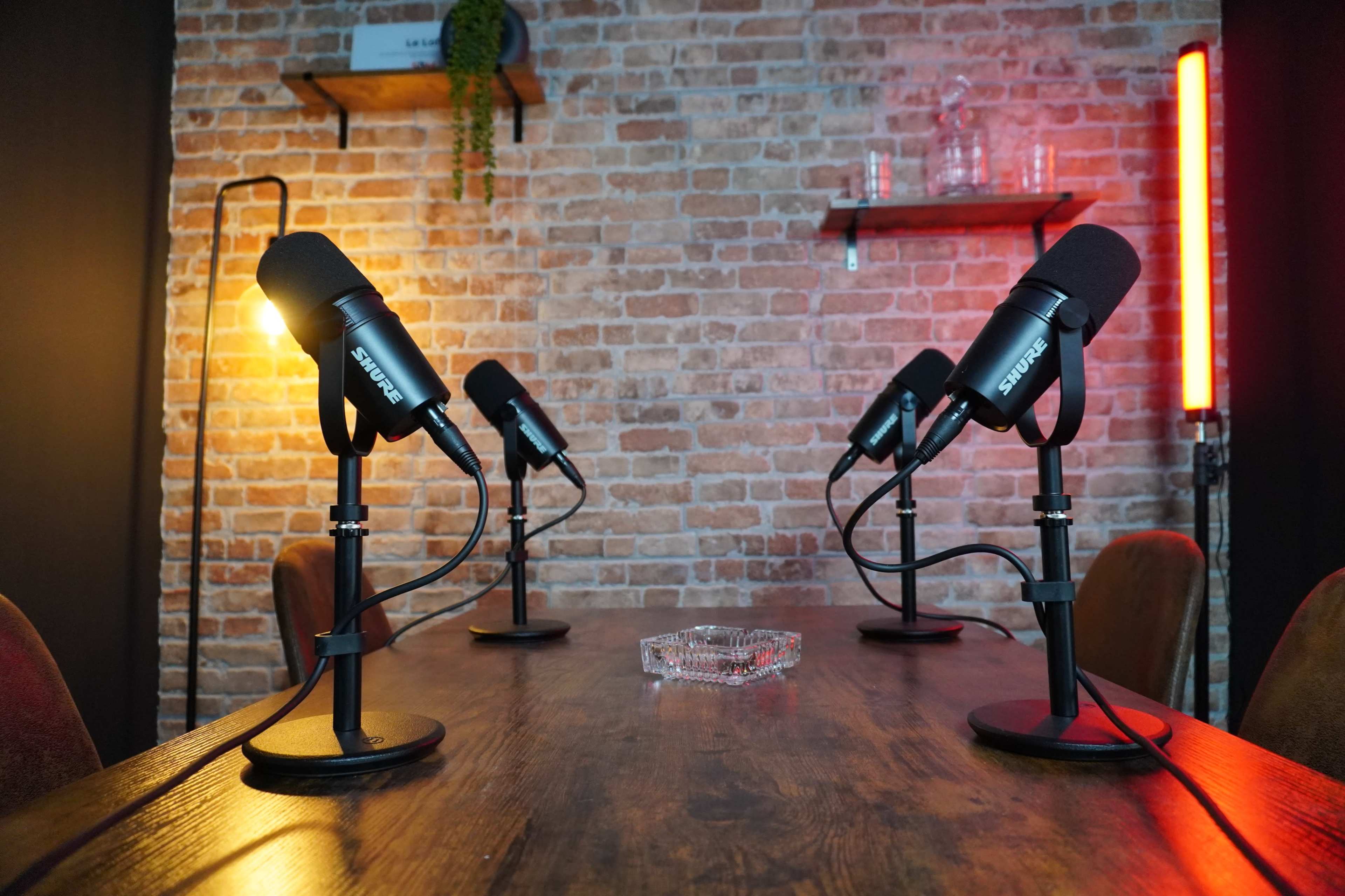 The image shows a wooden table set up with four black microphones facing each other in a room with exposed brick walls and minimalistic decor.