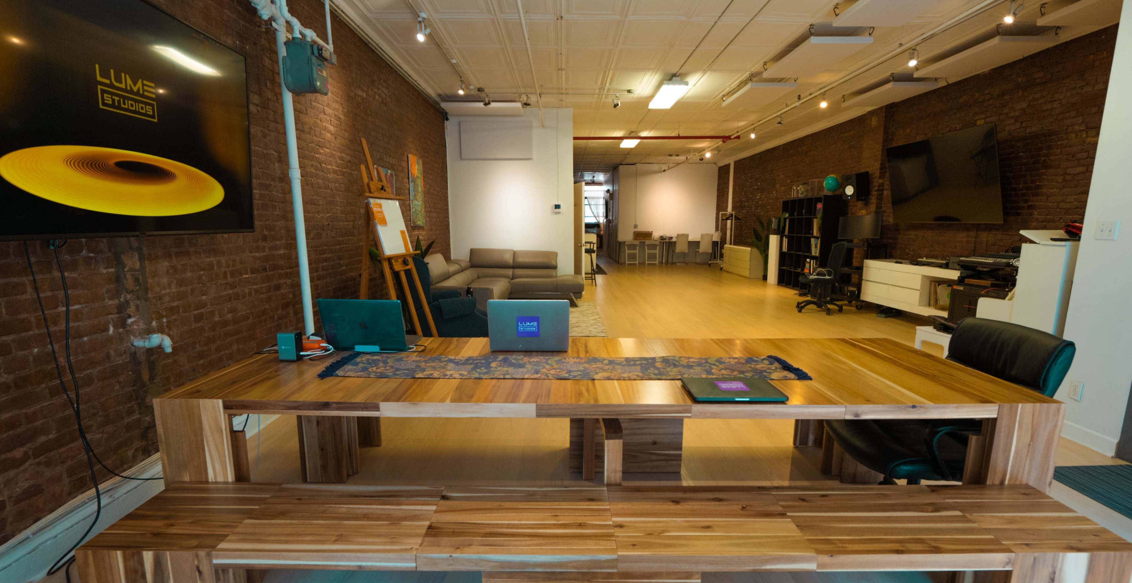 A spacious, modern studio features a large wooden table, a sleek laptop on it, and exposed brick walls, with seating and various workspaces in the background.