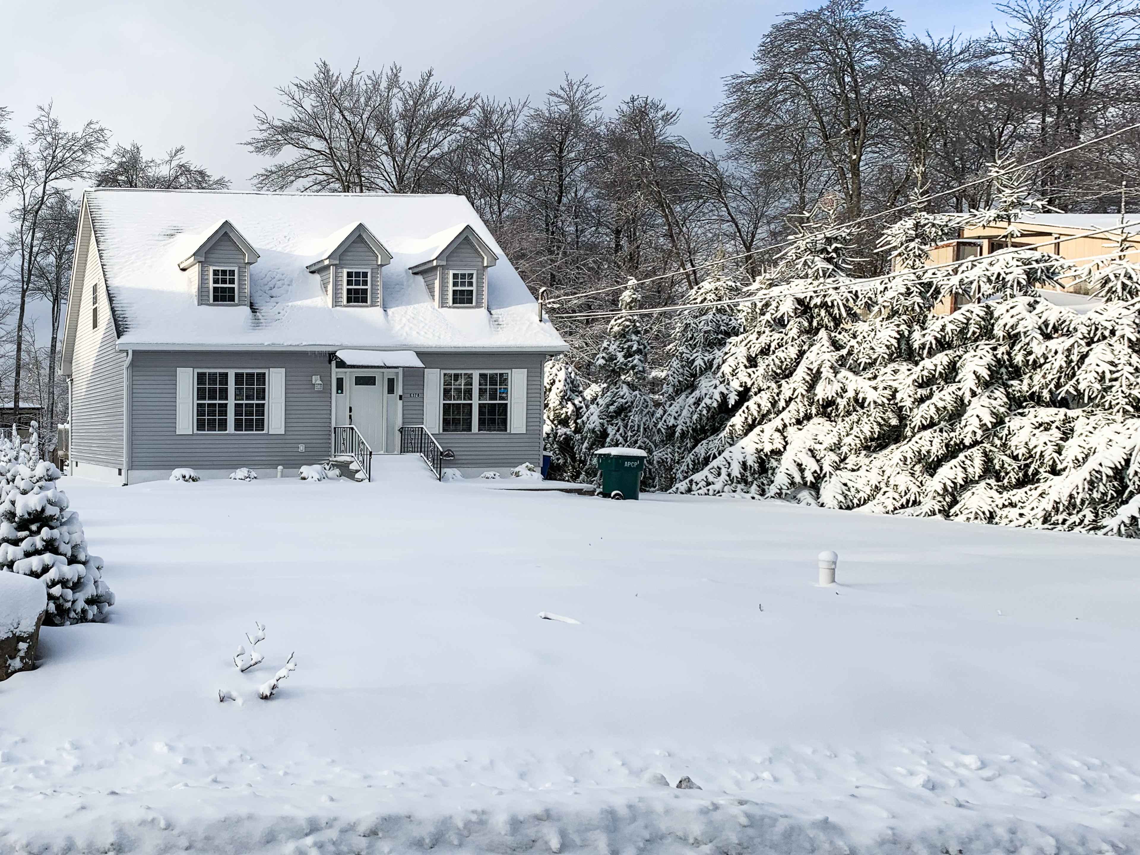 A two-story gray house with a steep roof sits in a snowy landscape, surrounded by tall trees and a green trash bin.