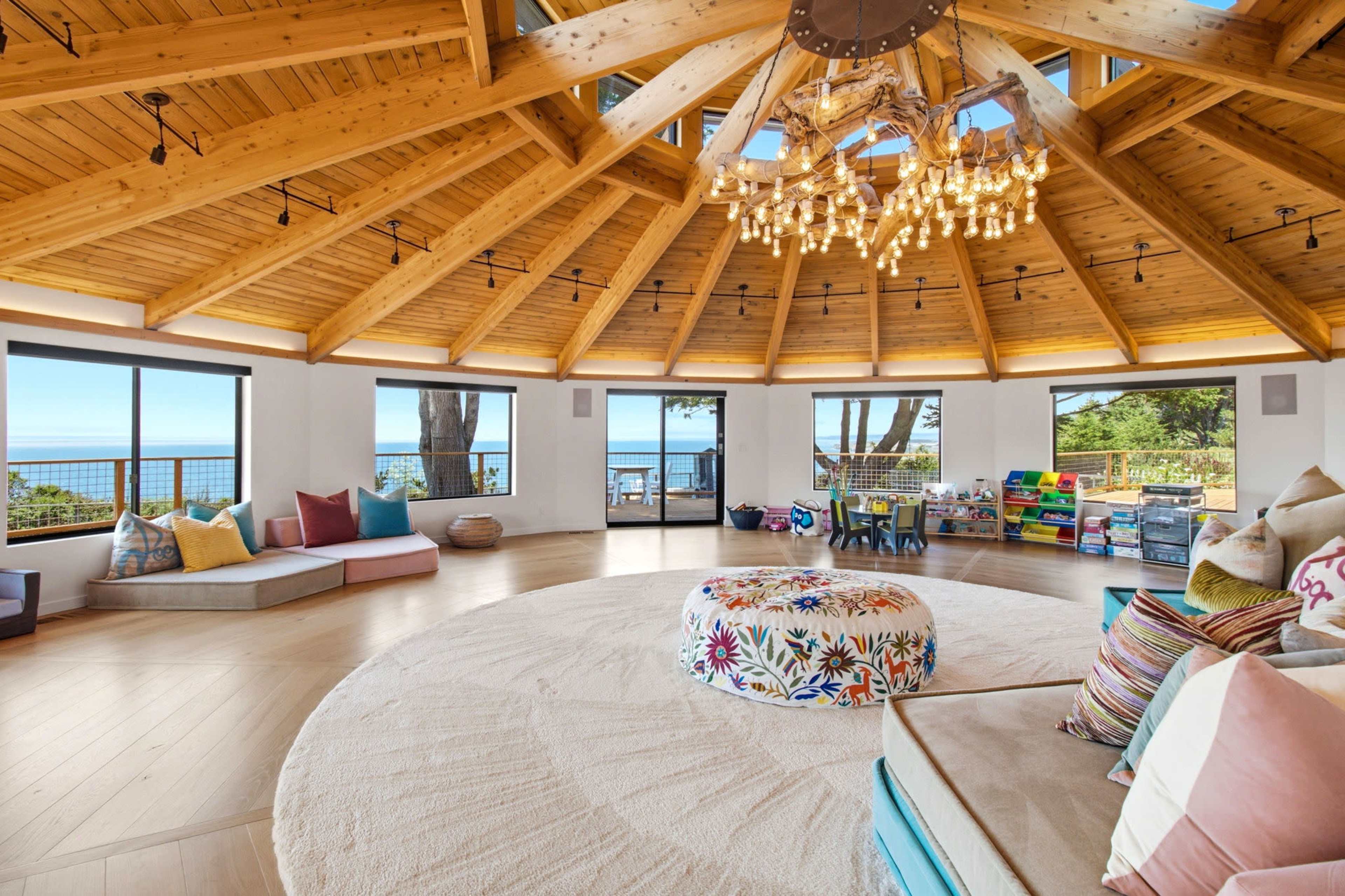 The image shows a spacious, circular room with a high wooden ceiling, large windows providing views of the ocean, and a cozy seating area with colorful cushions and a decorative pouf.