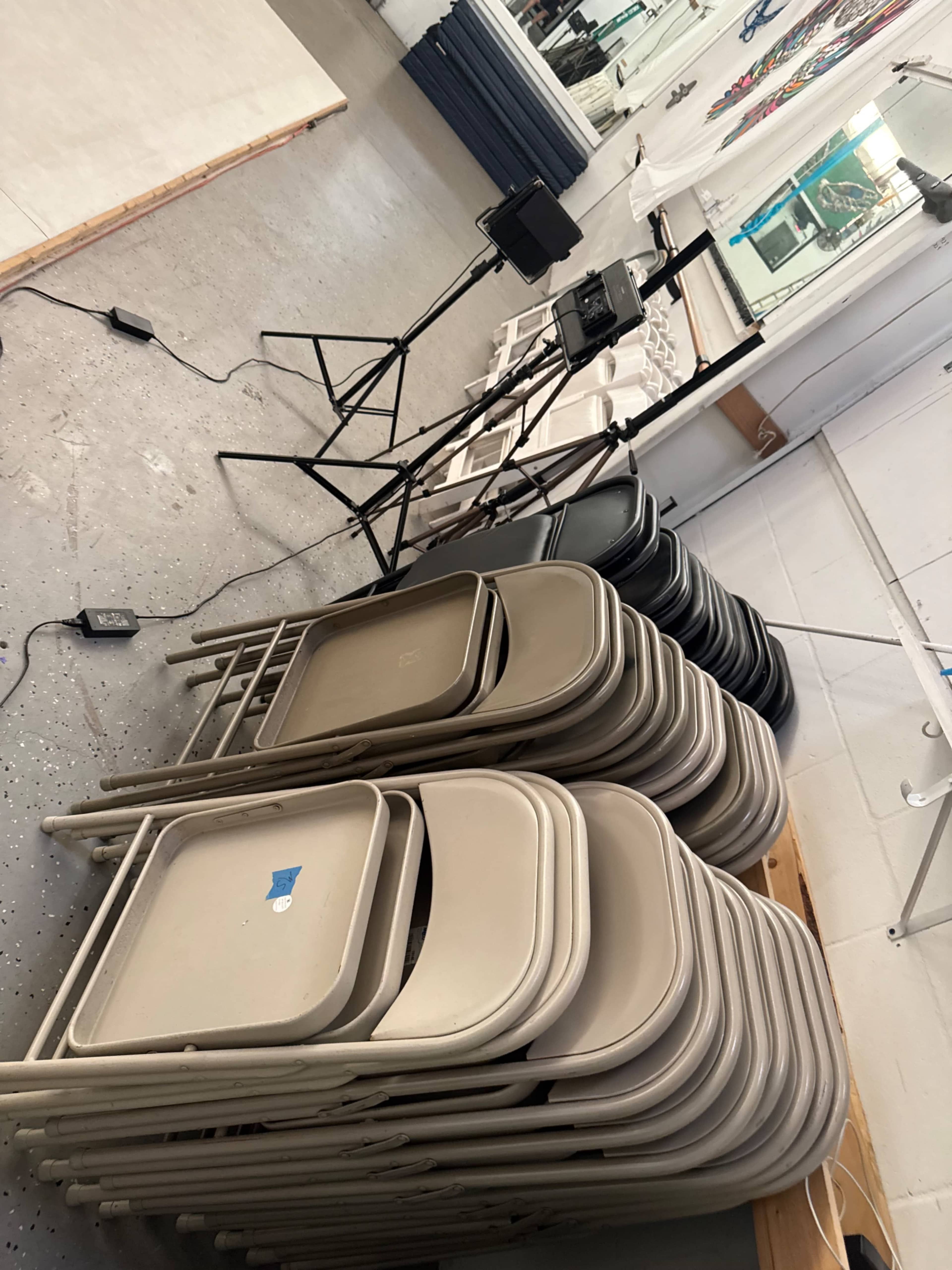 The image shows a stack of beige folding chairs next to a tripod with a camera set up in a well-lit indoor space.