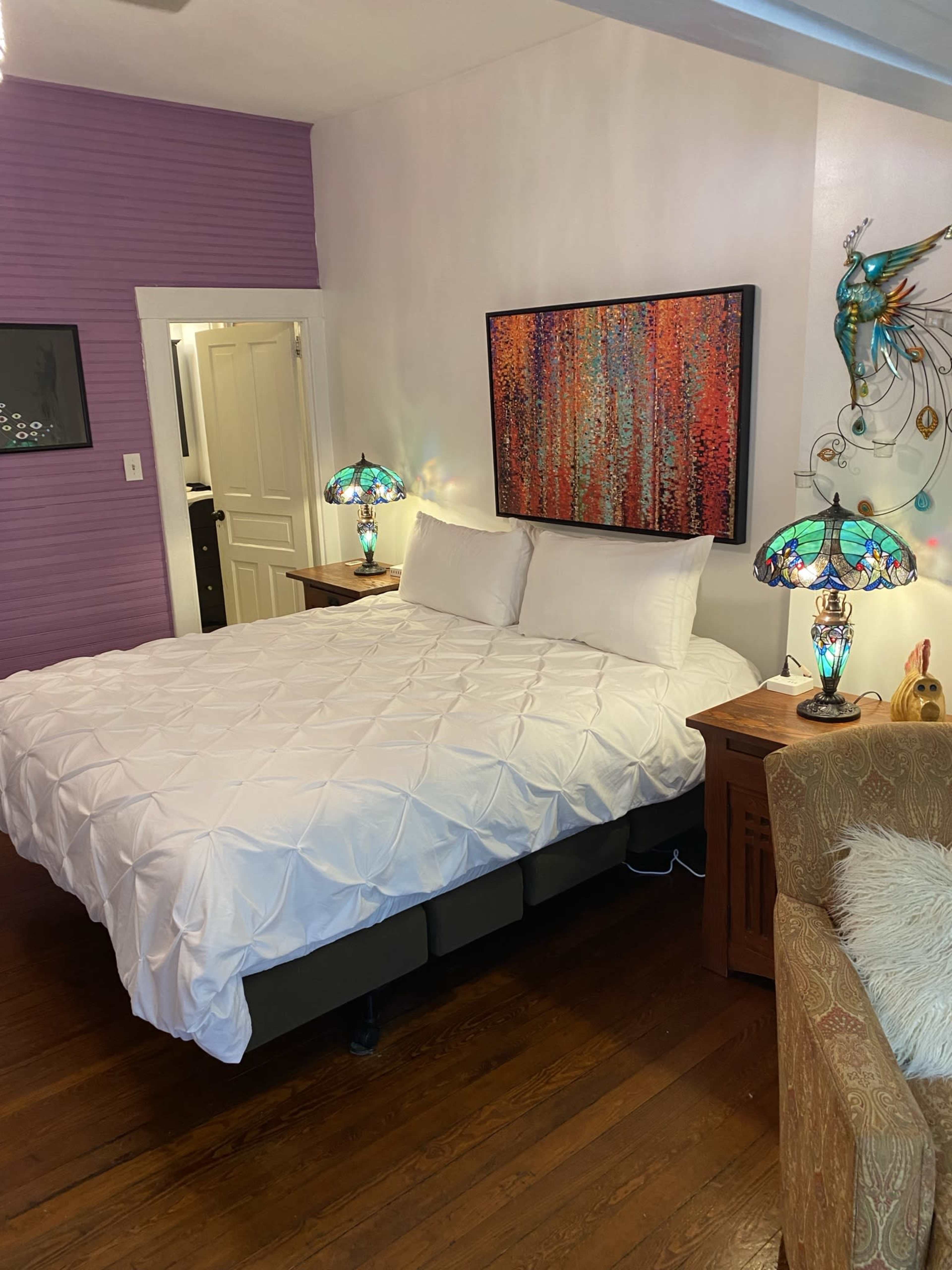 A cozy bedroom with a white bedspread, colorful artwork on the wall, and two ornate lamps on bedside tables.