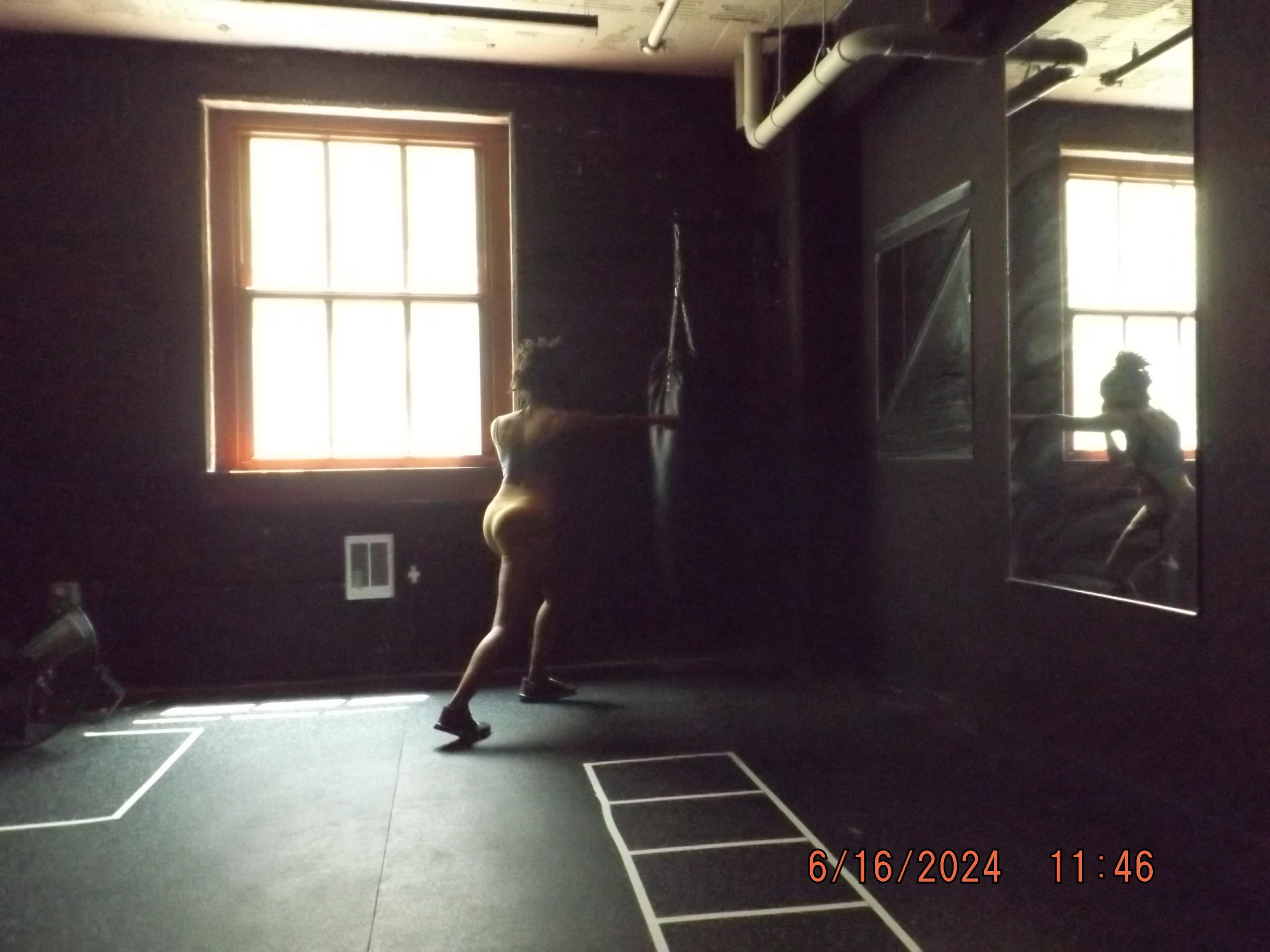 A person is hitting a punching bag in a dimly lit gym with large windows and a mirror reflecting their movement.