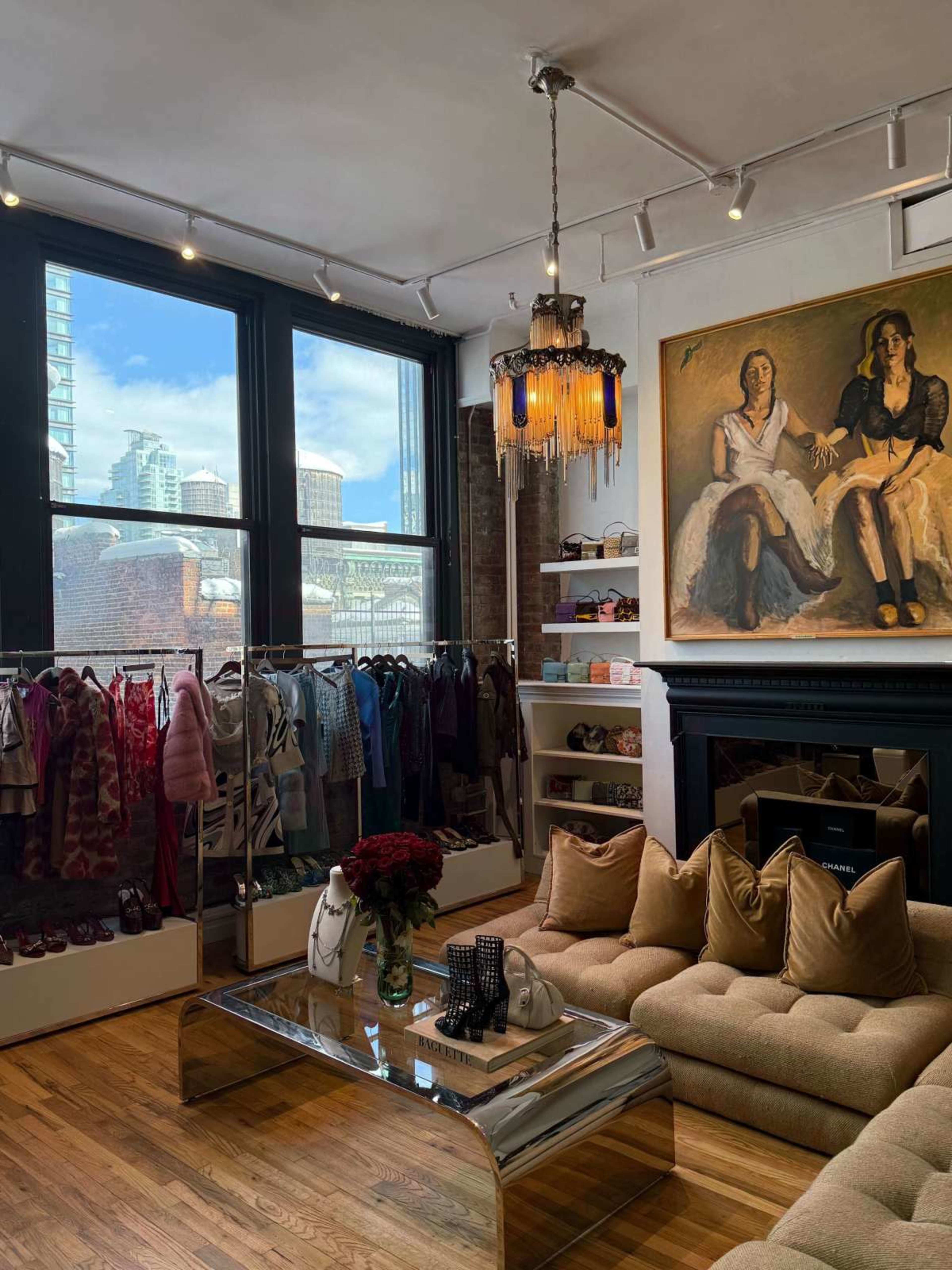 The image shows a stylish boutique with a cozy seating area, clothing racks, a large window displaying a cityscape, and a notable chandelier hanging from the ceiling.