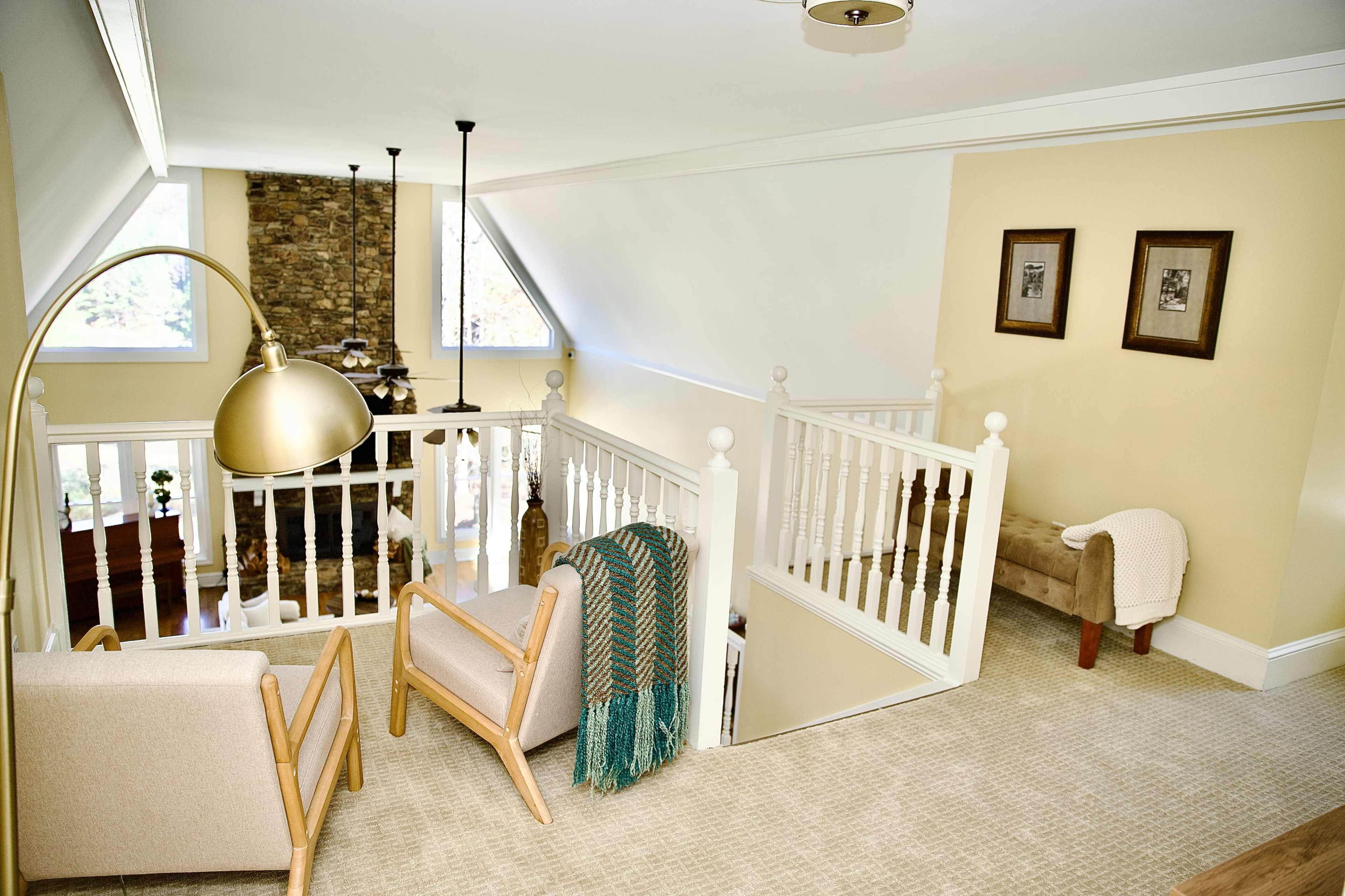 The image shows an airy loft space featuring two armchairs, a floor lamp, and a view of a stone fireplace below.