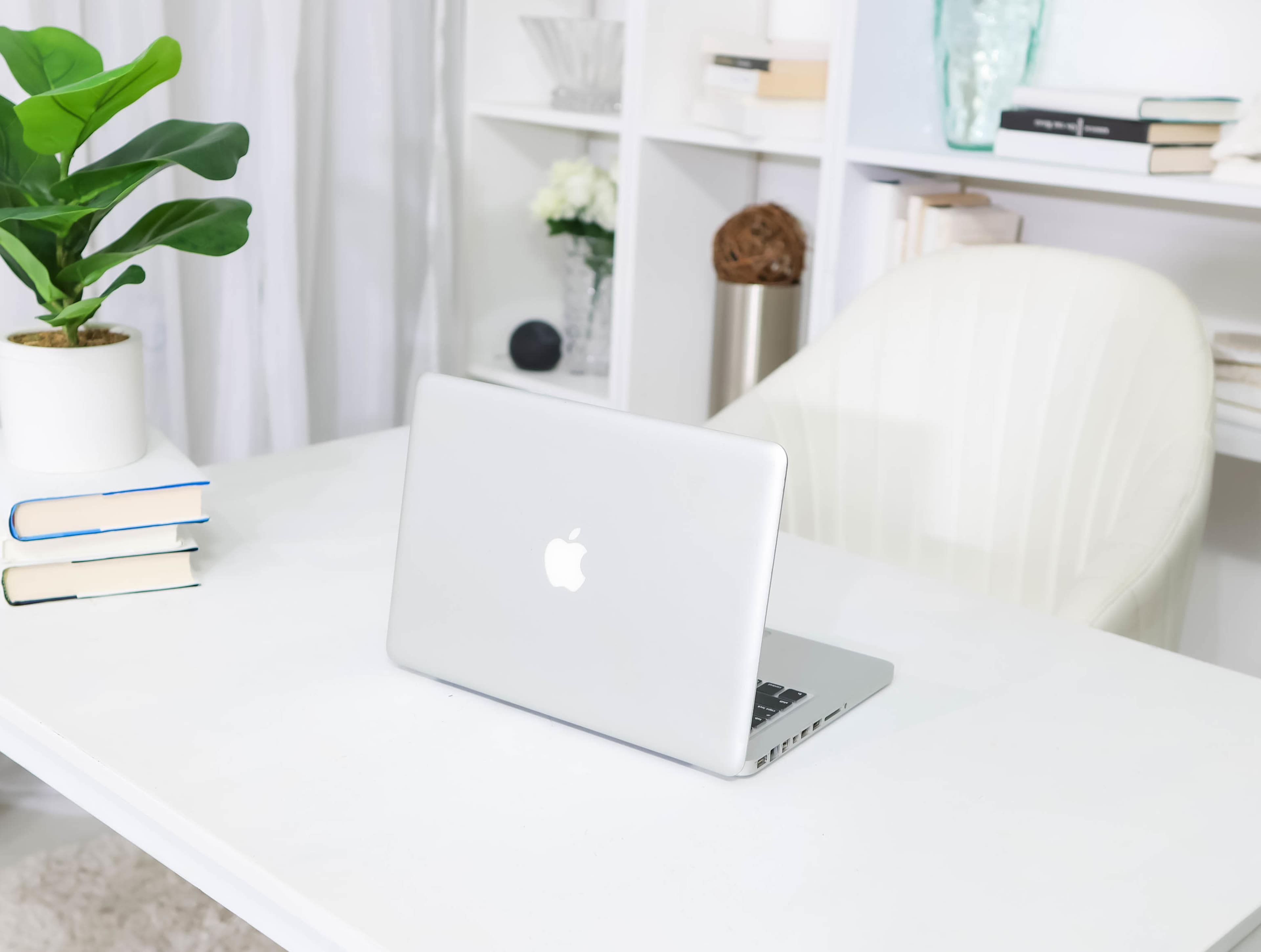 A silver laptop is open on a white desk with books stacked beside a potted plant in a well-organized room.