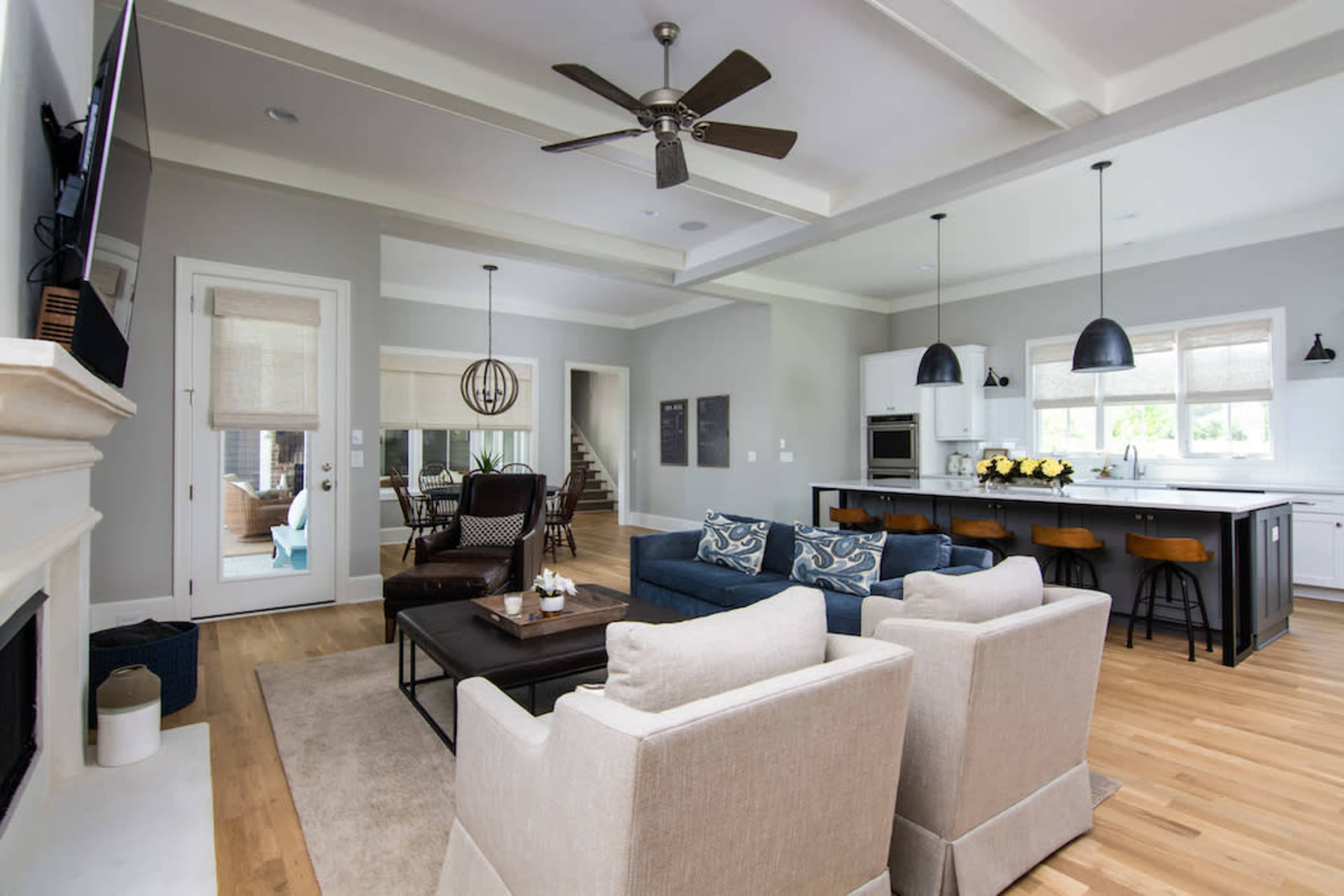 The image shows a modern living room with a blue couch, cream-colored armchairs, and a kitchen area featuring dark cabinetry and pendant lights.