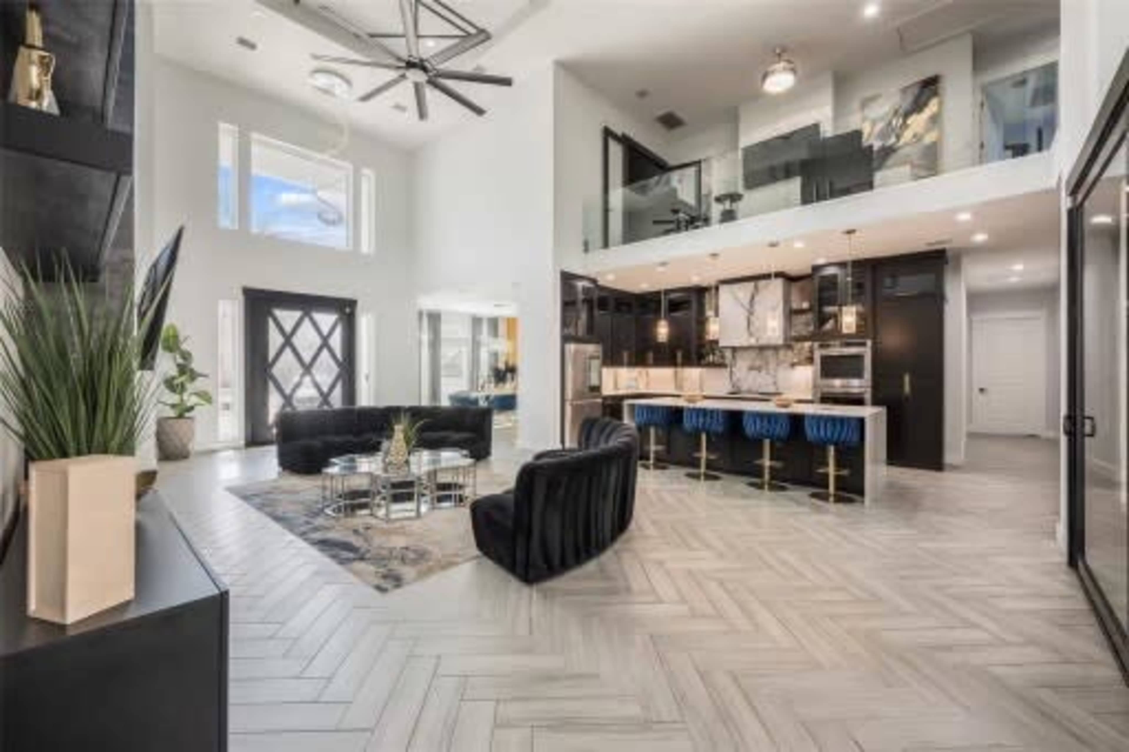 The image shows a modern living room with tall ceilings, featuring black furniture, a patterned rug, and a kitchen area with a large island and bar seating.