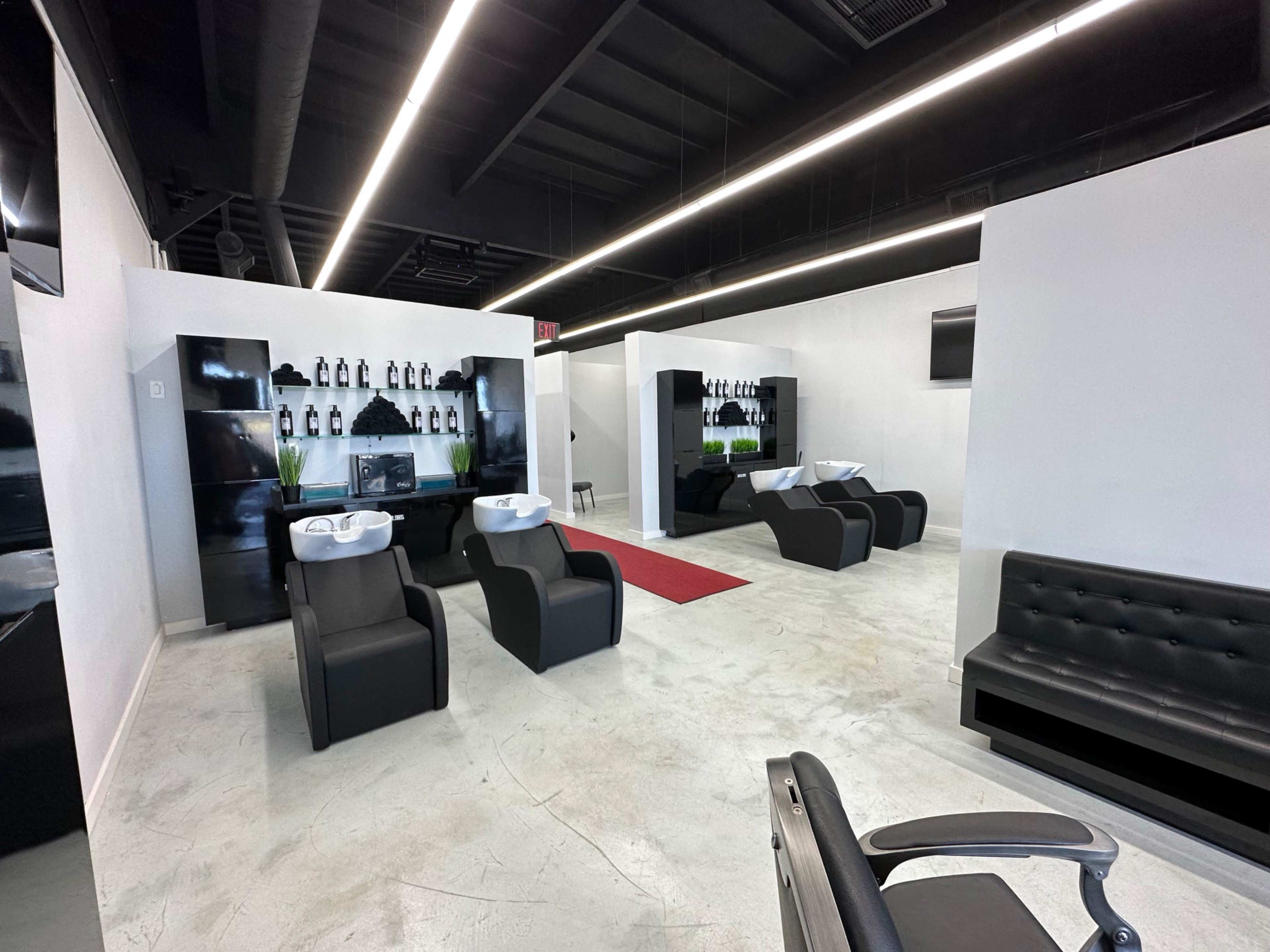 The image shows a modern salon interior featuring black styling chairs, wash stations, a red carpet, and a minimalist design with white walls and overhead lighting.