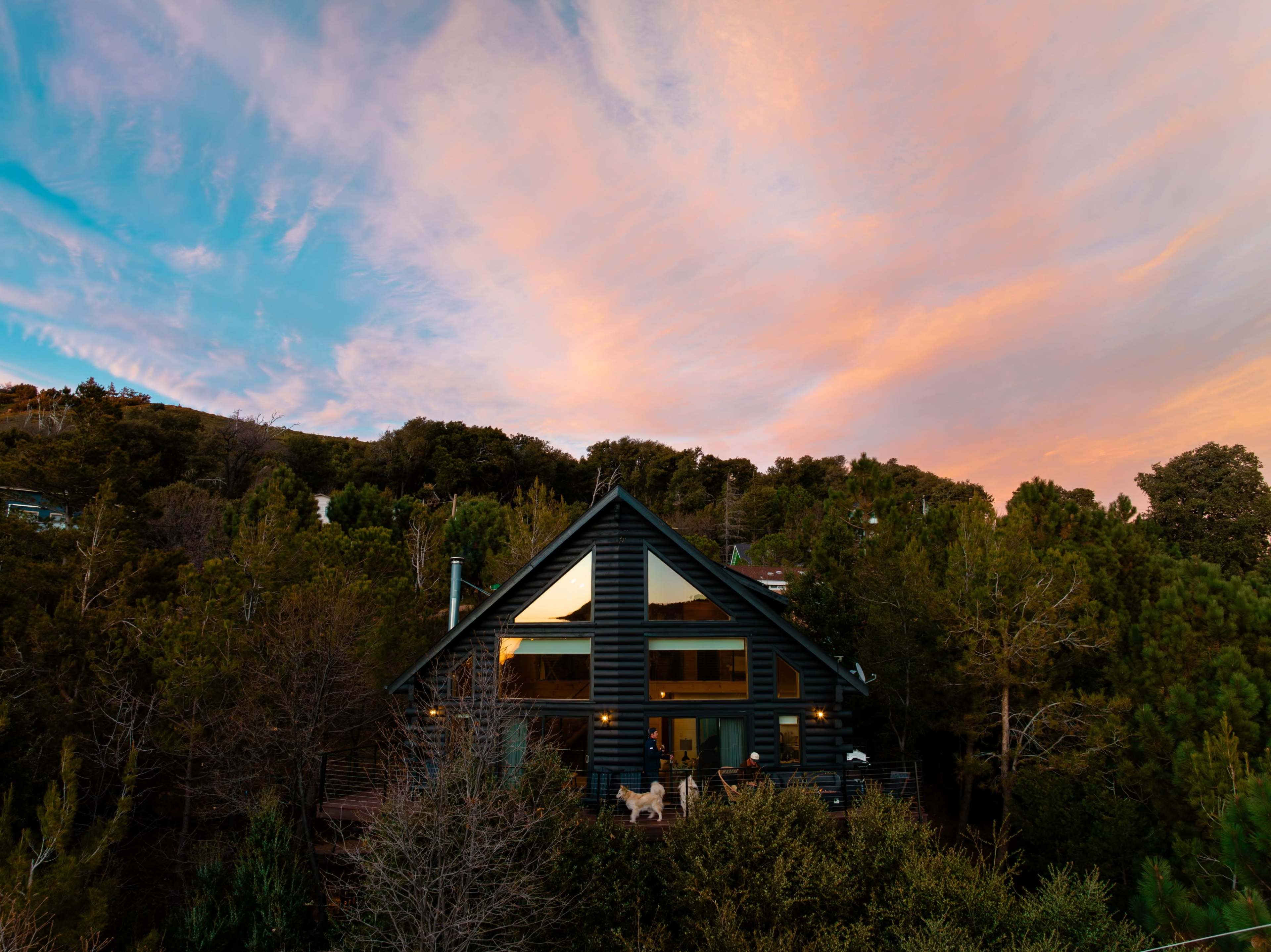A wooden cabin surrounded by trees sits beneath a colorful sunset sky.