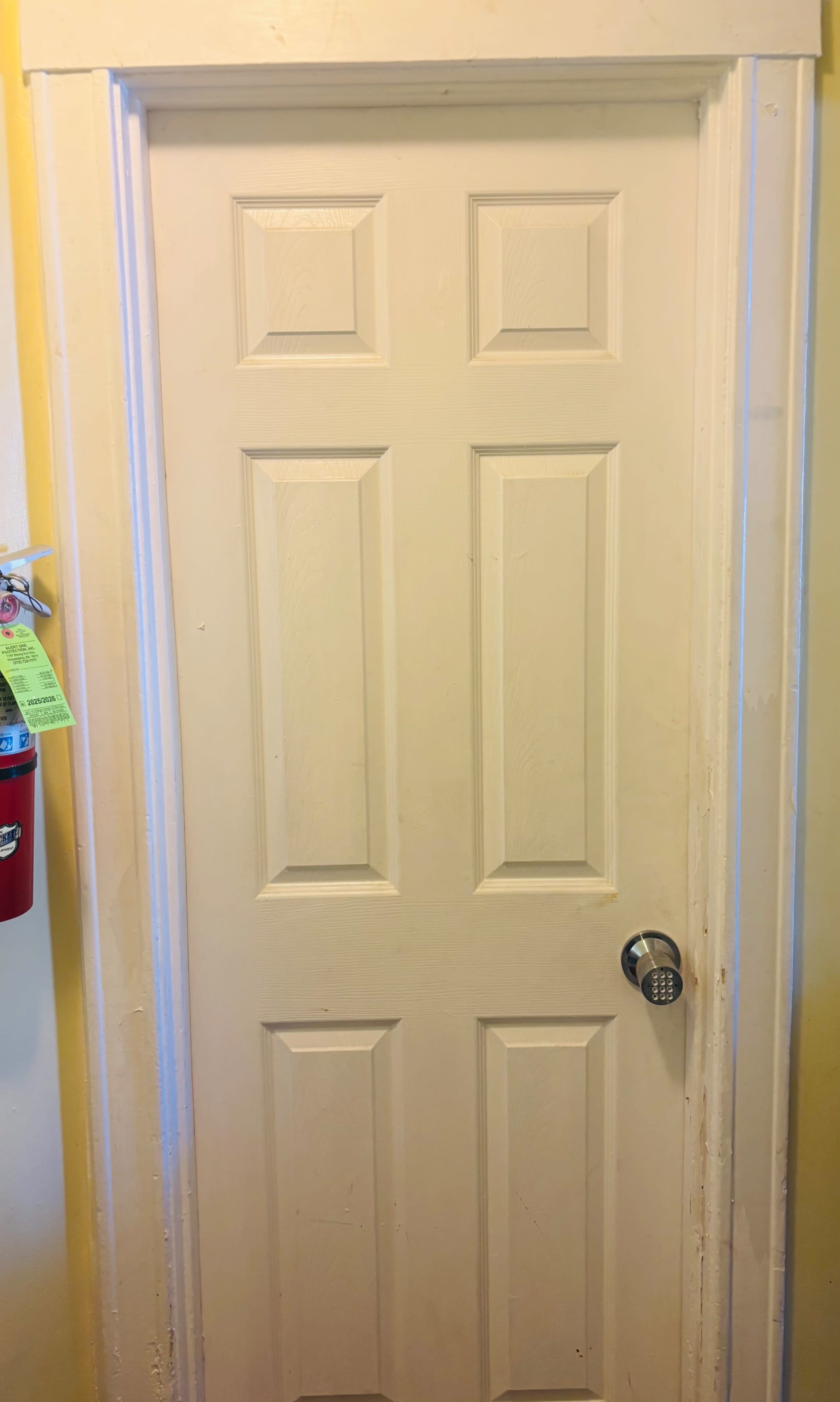 The image shows a closed, six-panel white door with a round silver doorknob and a combination lock, set against a yellow wall.