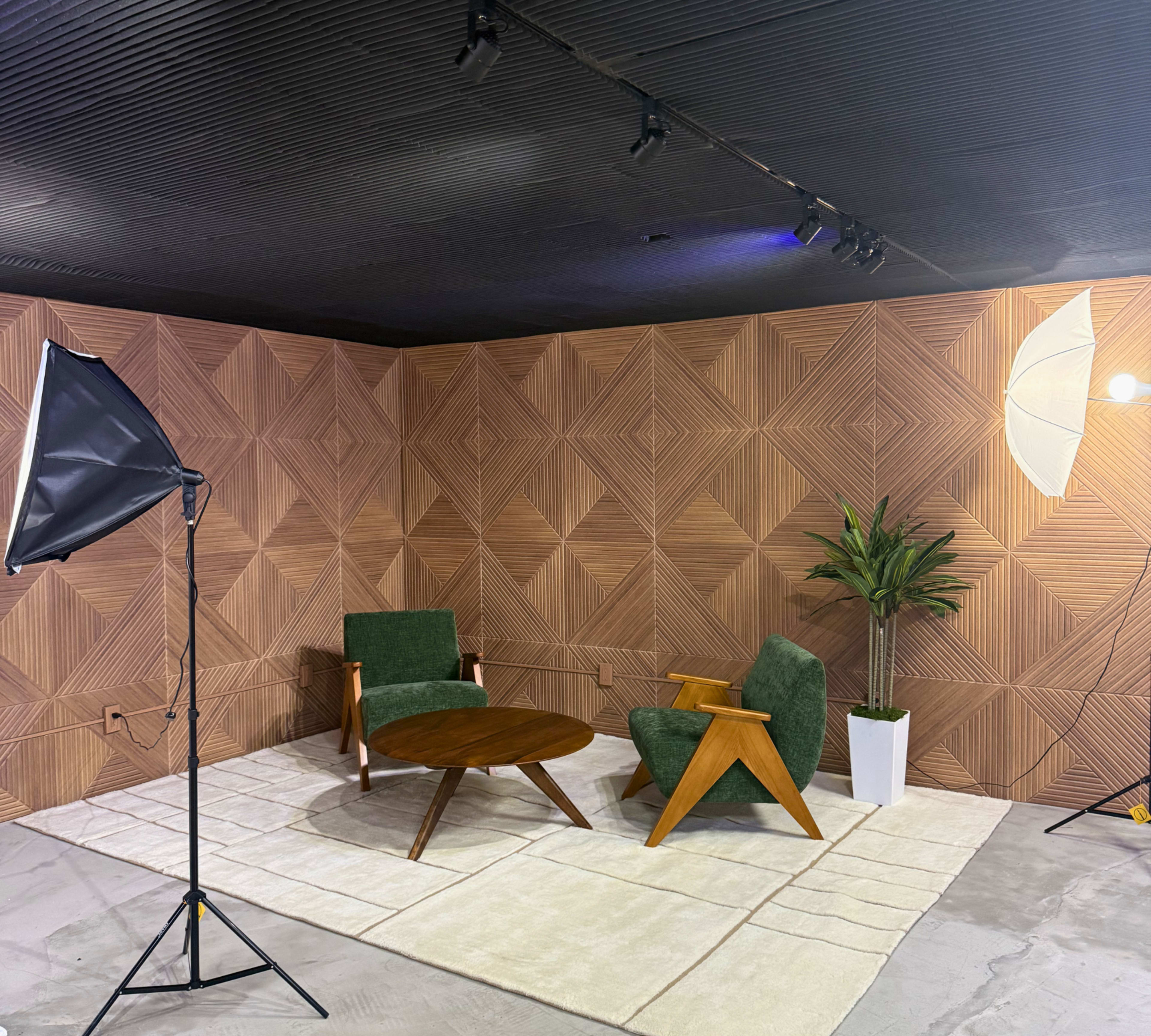 The image shows a modern interior space featuring a geometric wood-paneled wall, two green armchairs, a round wooden table, a potted plant, and soft lighting equipment.