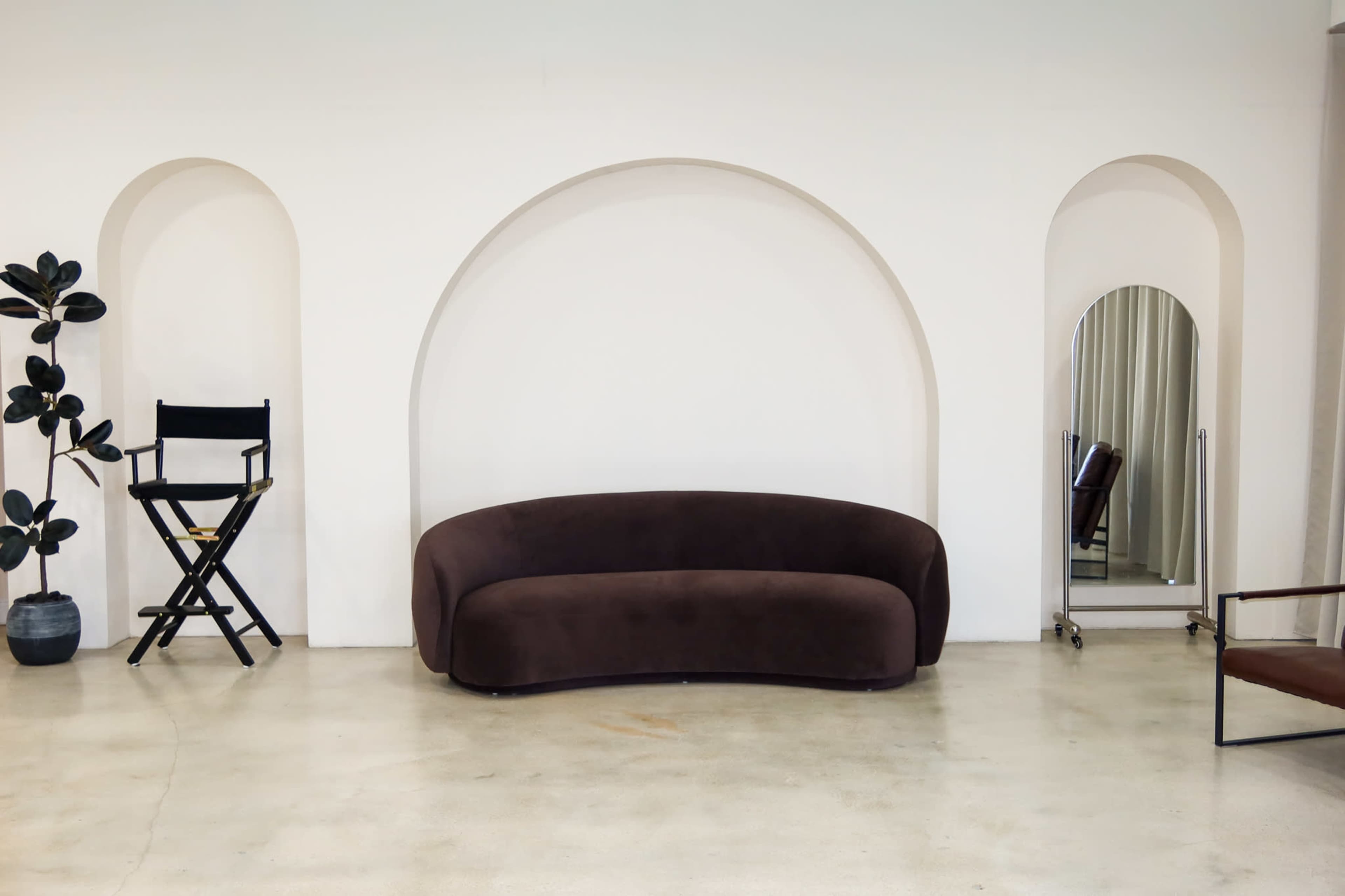 The image shows a minimalist interior featuring a brown curved sofa, a director's chair, a full-length mirror, and a potted plant against a white wall with arched niches.