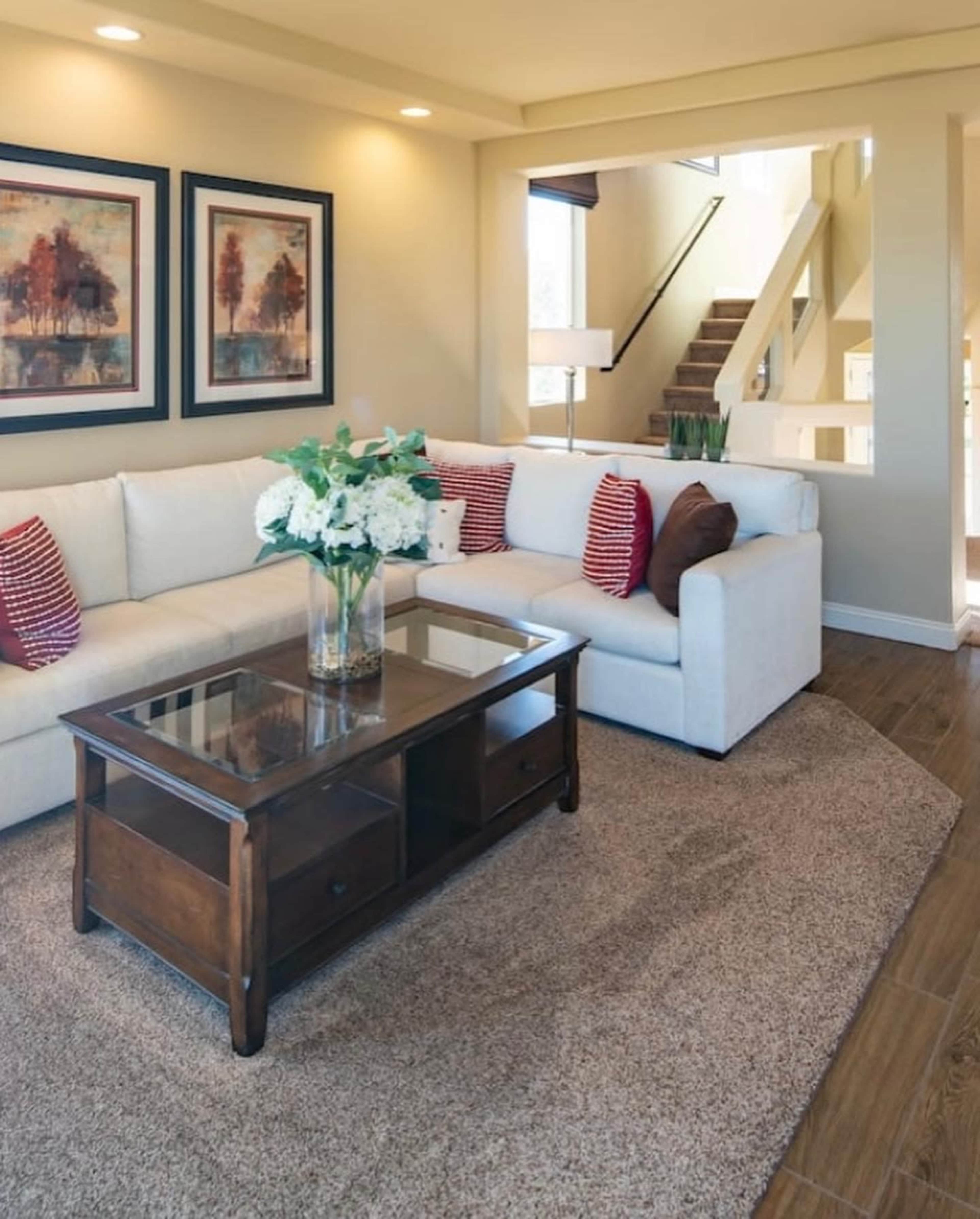 The image shows a neatly arranged living room with a light-colored sectional sofa, a wooden coffee table, and a bouquet of flowers on top, accompanied by framed artwork on the walls.