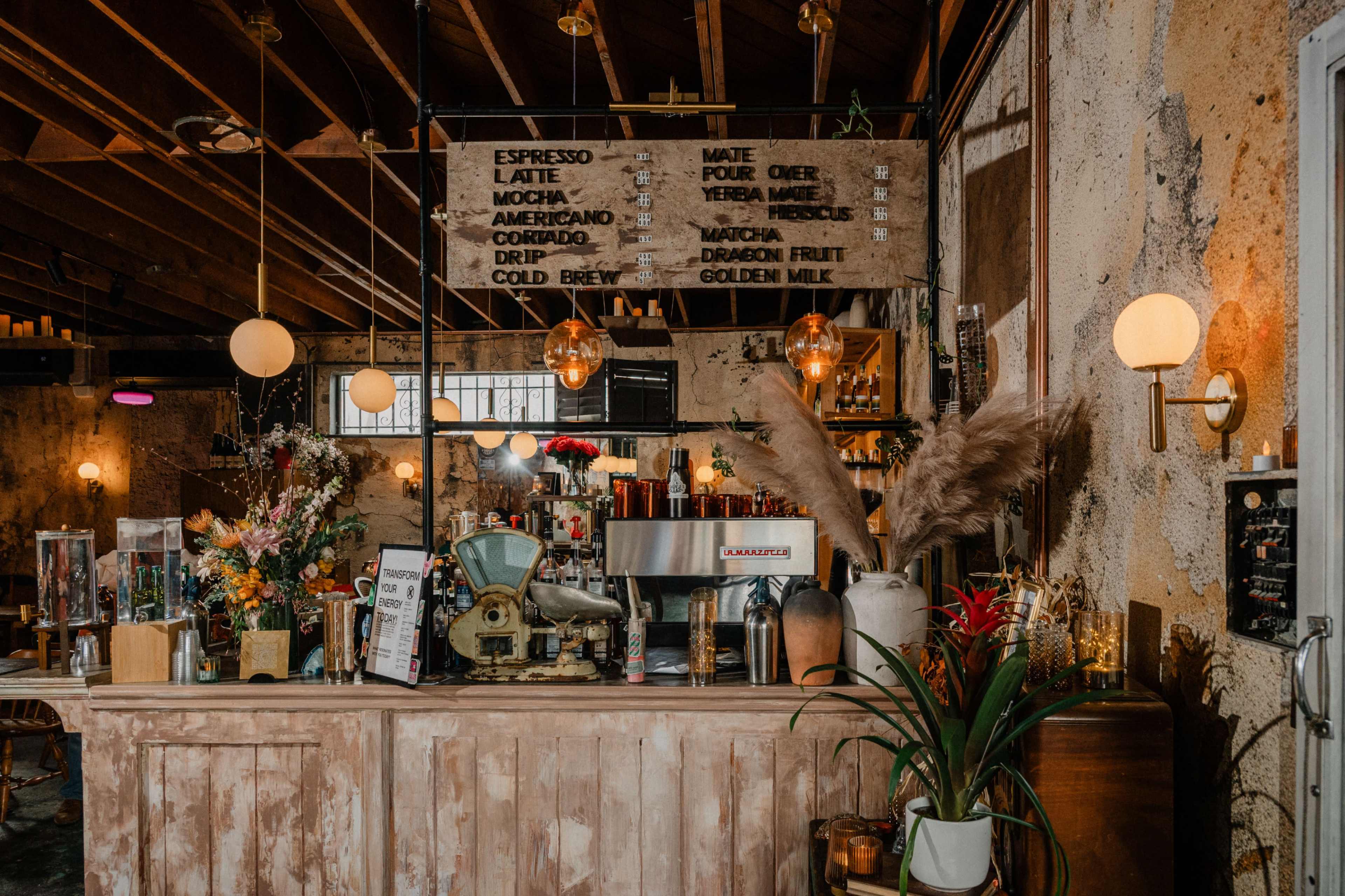 A rustic café counter features a coffee machine and a menu board with various beverage options, surrounded by decorative plants and flowers.