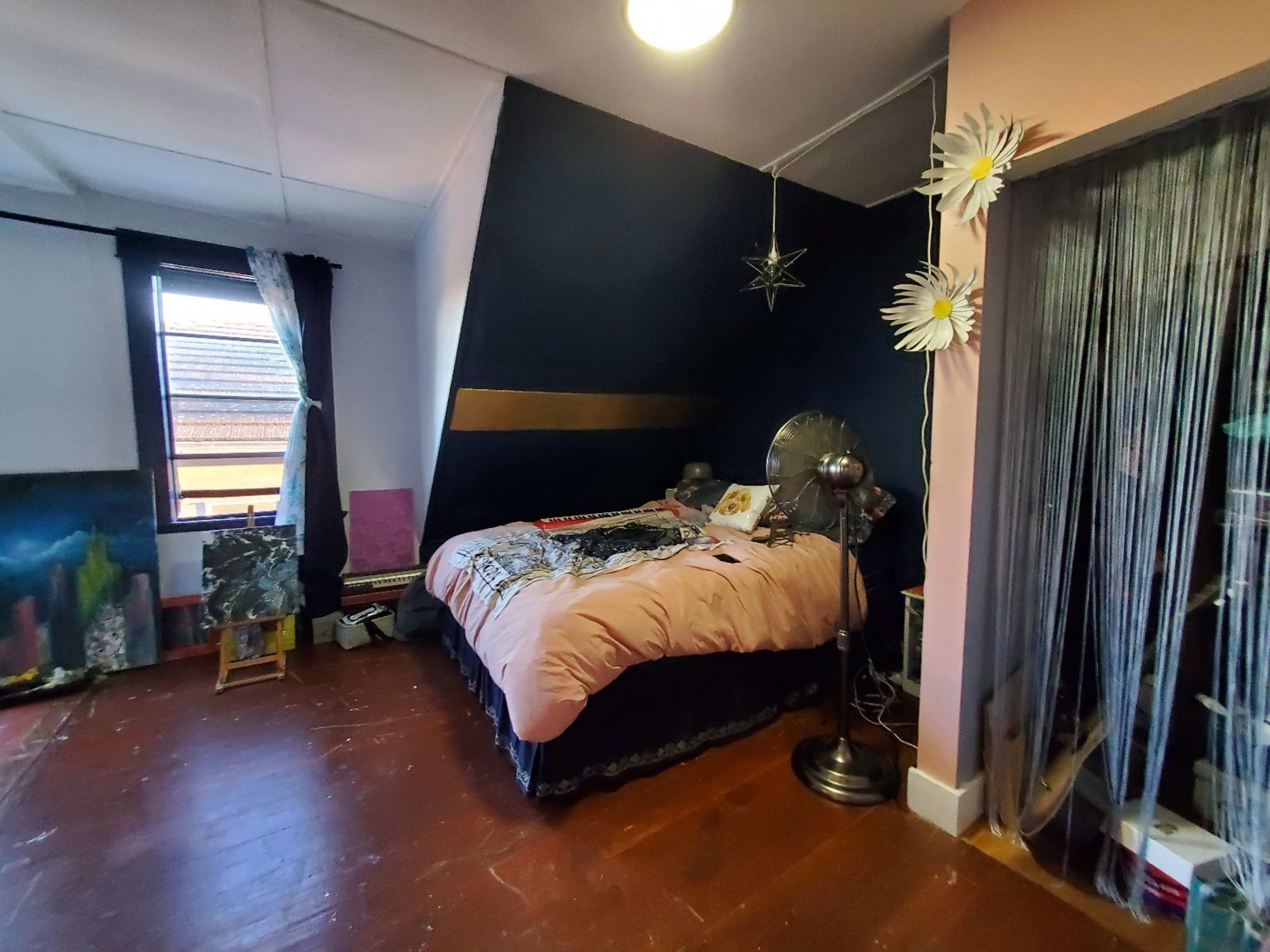 The bedroom features a bed with a pink comforter, a window with dark curtains, and a decorative fan beside it, along with paintings and wall decorations.