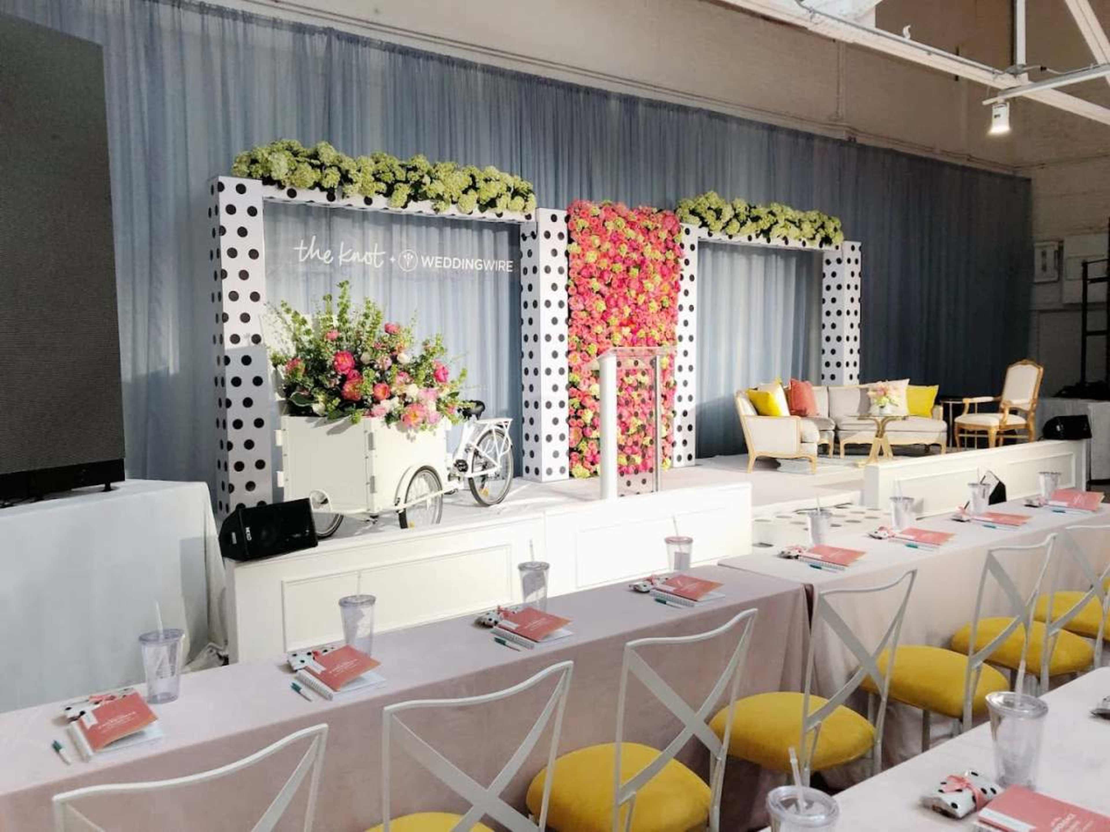 The image shows a decorated event space with a floral backdrop, white stage platform, and tables arranged in front with place settings.