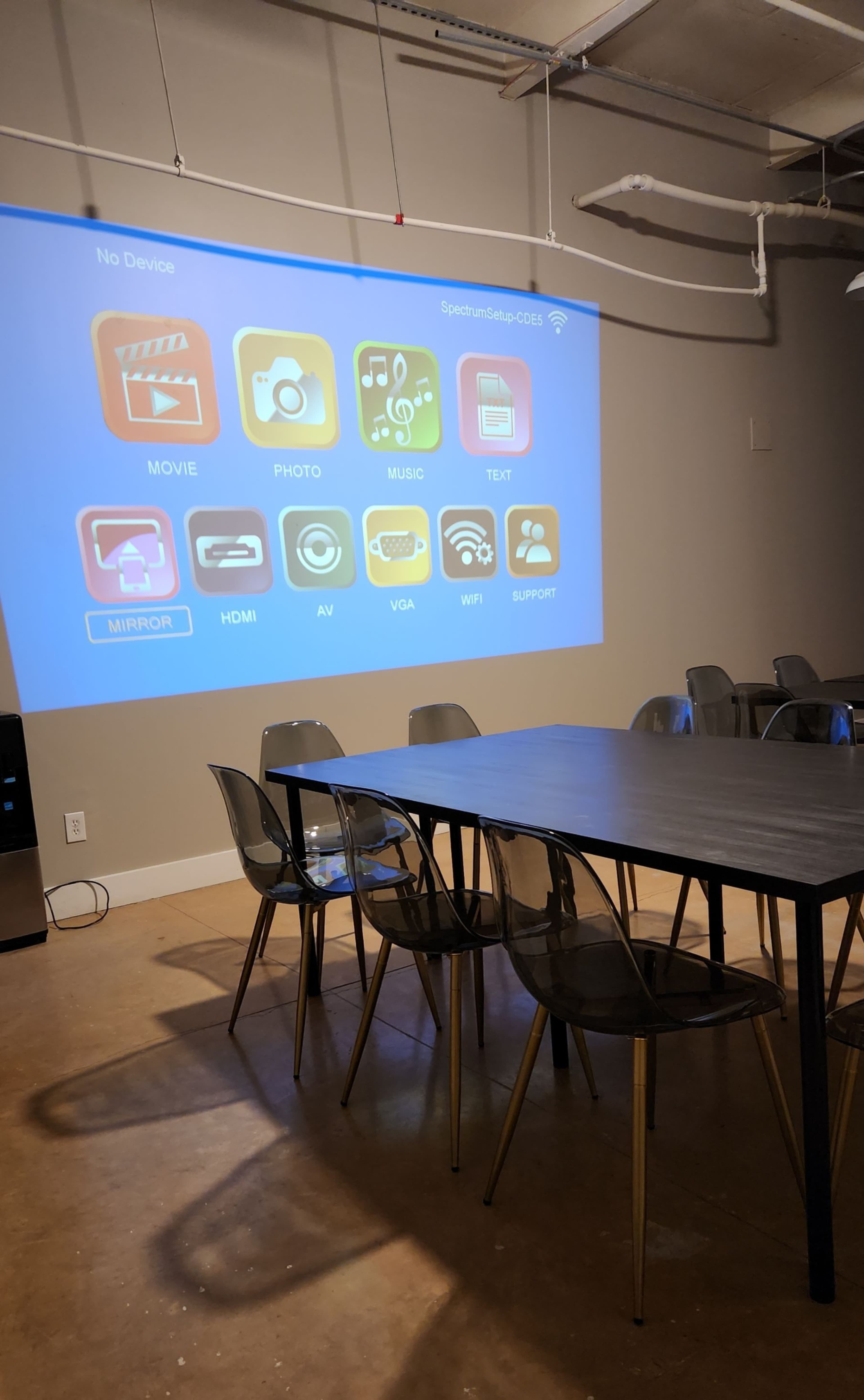 A conference room with a large projector screen displaying options for movie, photo, music, text, and device connections, set up around a long table and plastic chairs.