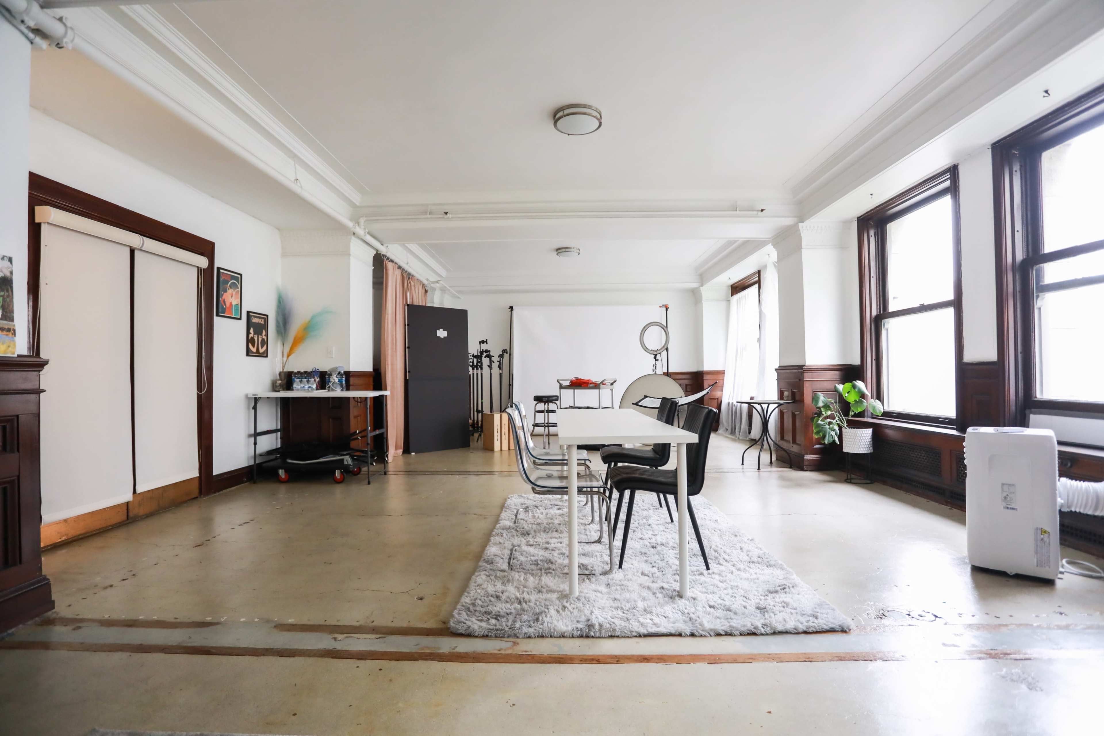 The image shows a spacious room with large windows, a dining table surrounded by chairs, and a light rug on a concrete floor.