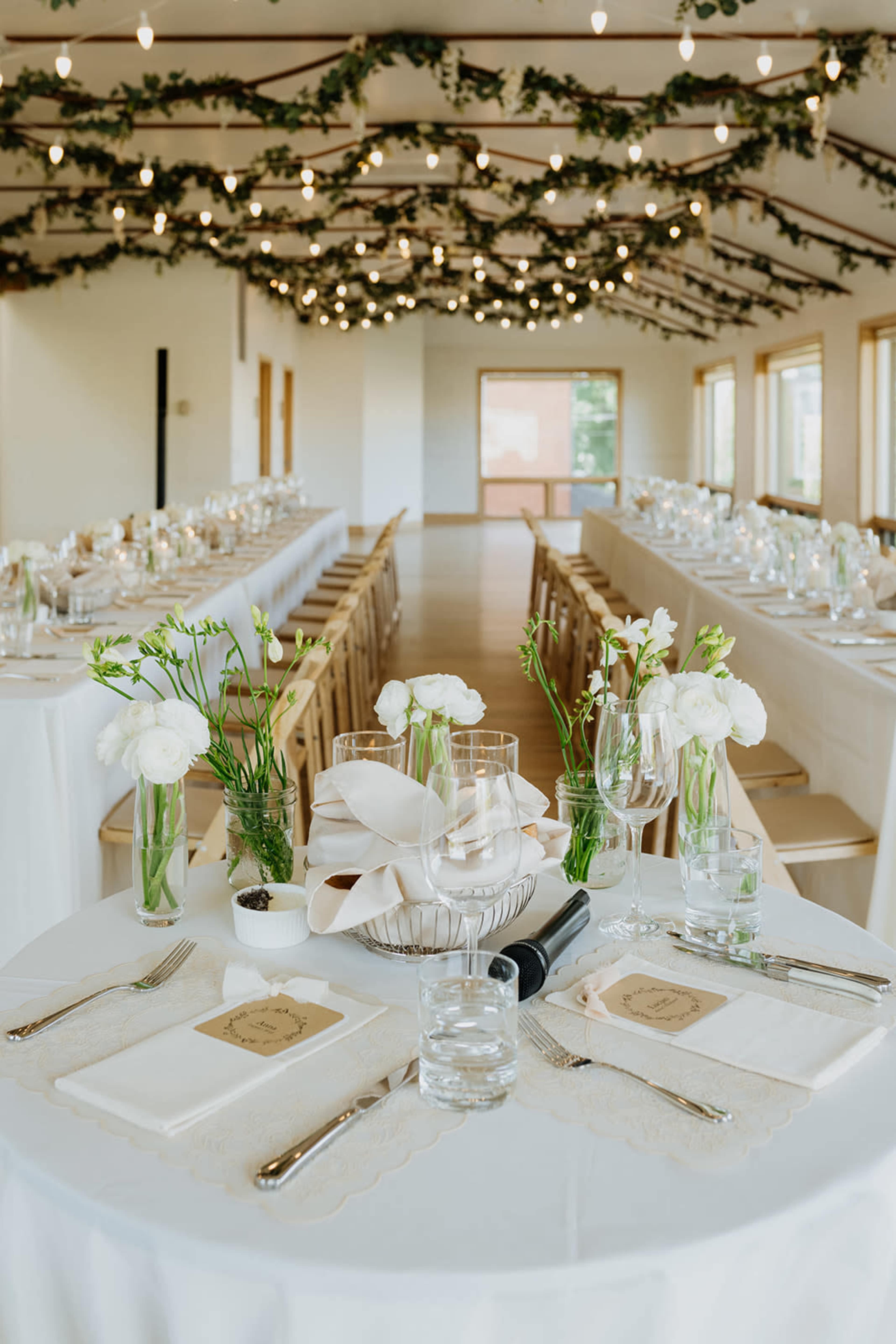 A beautifully arranged banquet hall features long tables with white linens, centerpieces of flowers and candles, surrounded by decorative greenery and soft lighting.