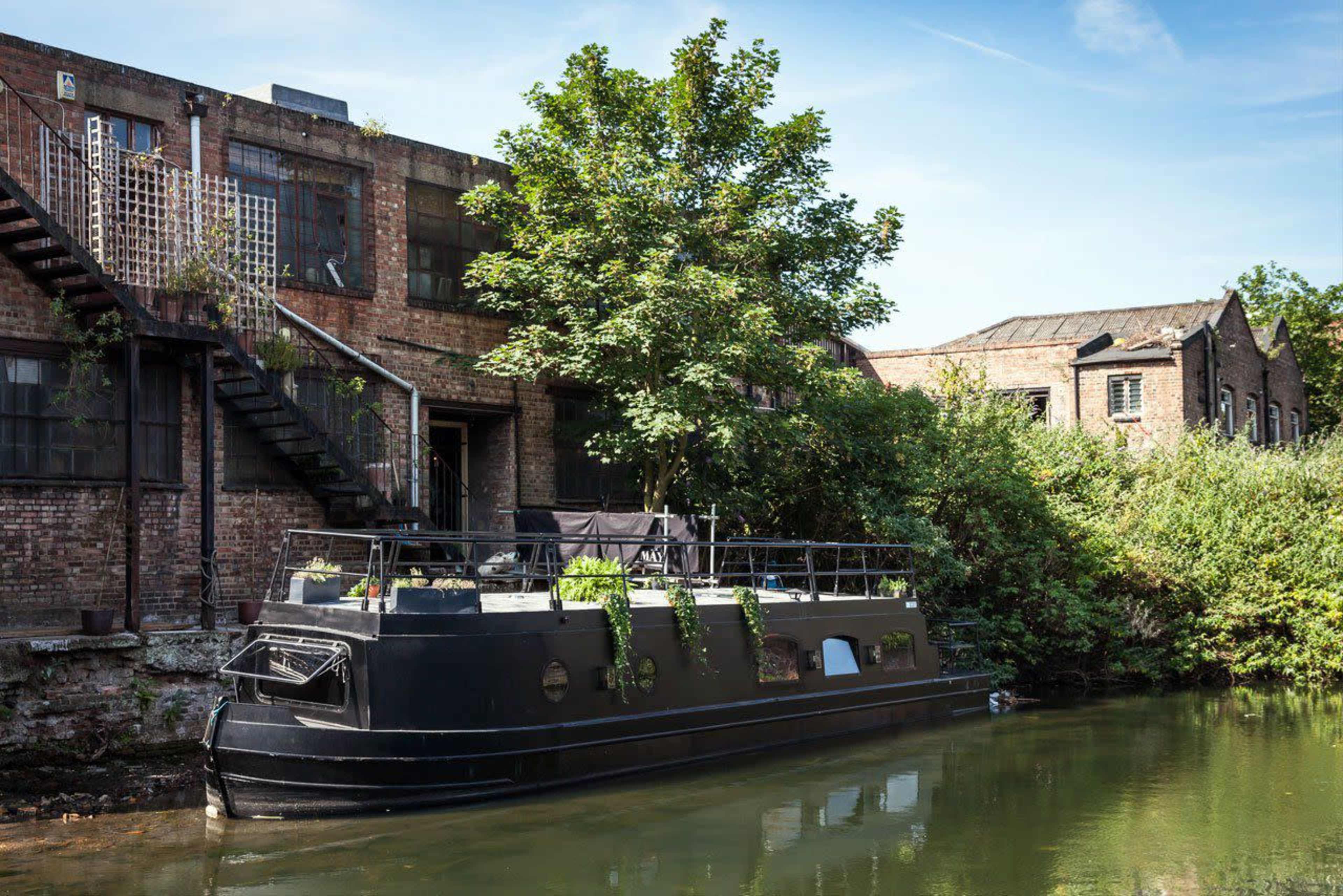 A black canal boat is moored next to a brick building with overgrown vegetation along the waterway.