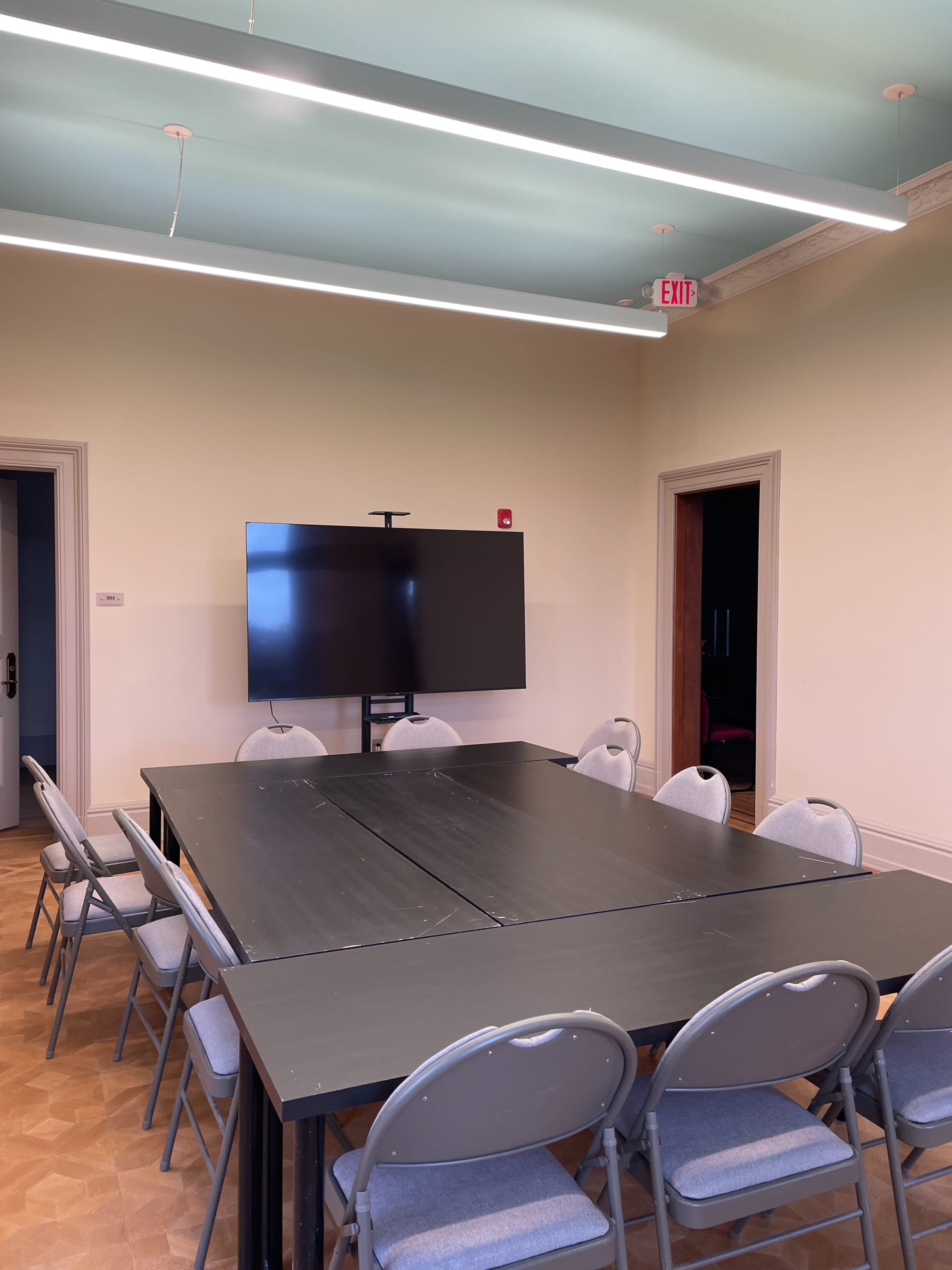 A modern conference room features a large table surrounded by gray chairs and a flat-screen monitor mounted on a stand.