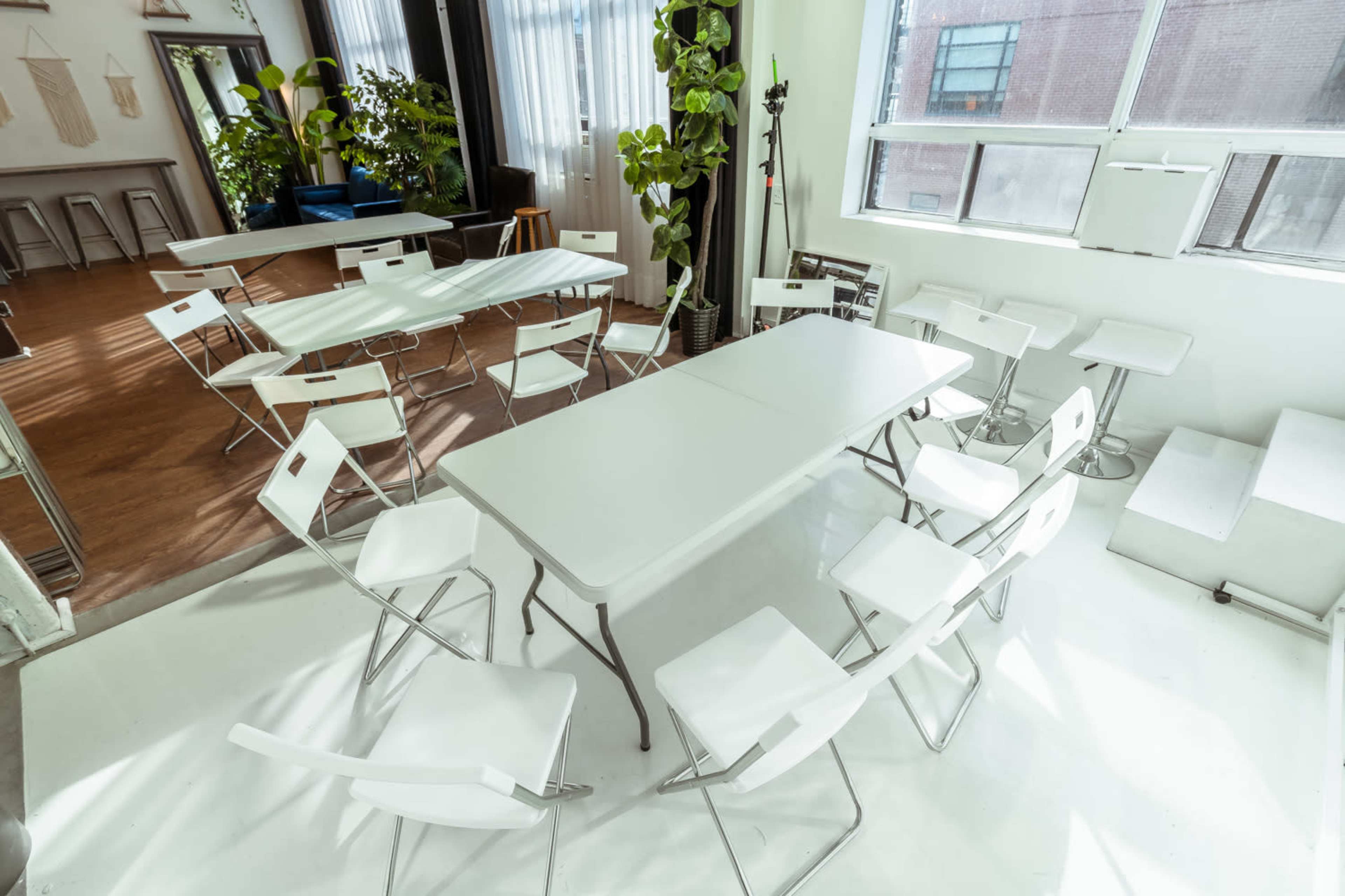 A bright room arranged with multiple folding tables and chairs, surrounded by large windows and greenery.