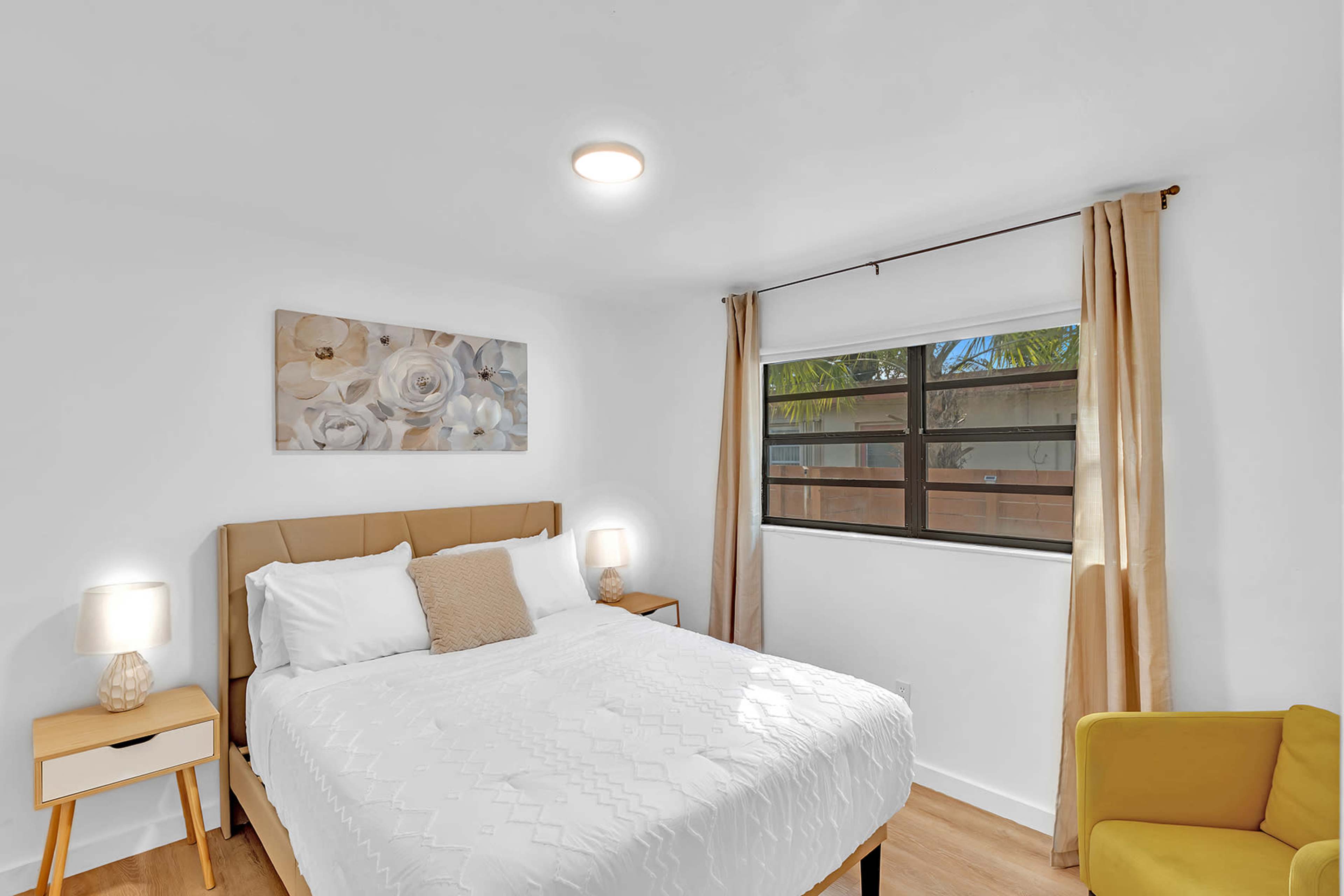 A bright bedroom featuring a queen-sized bed with white bedding, a framed floral artwork, and a window with beige curtains.