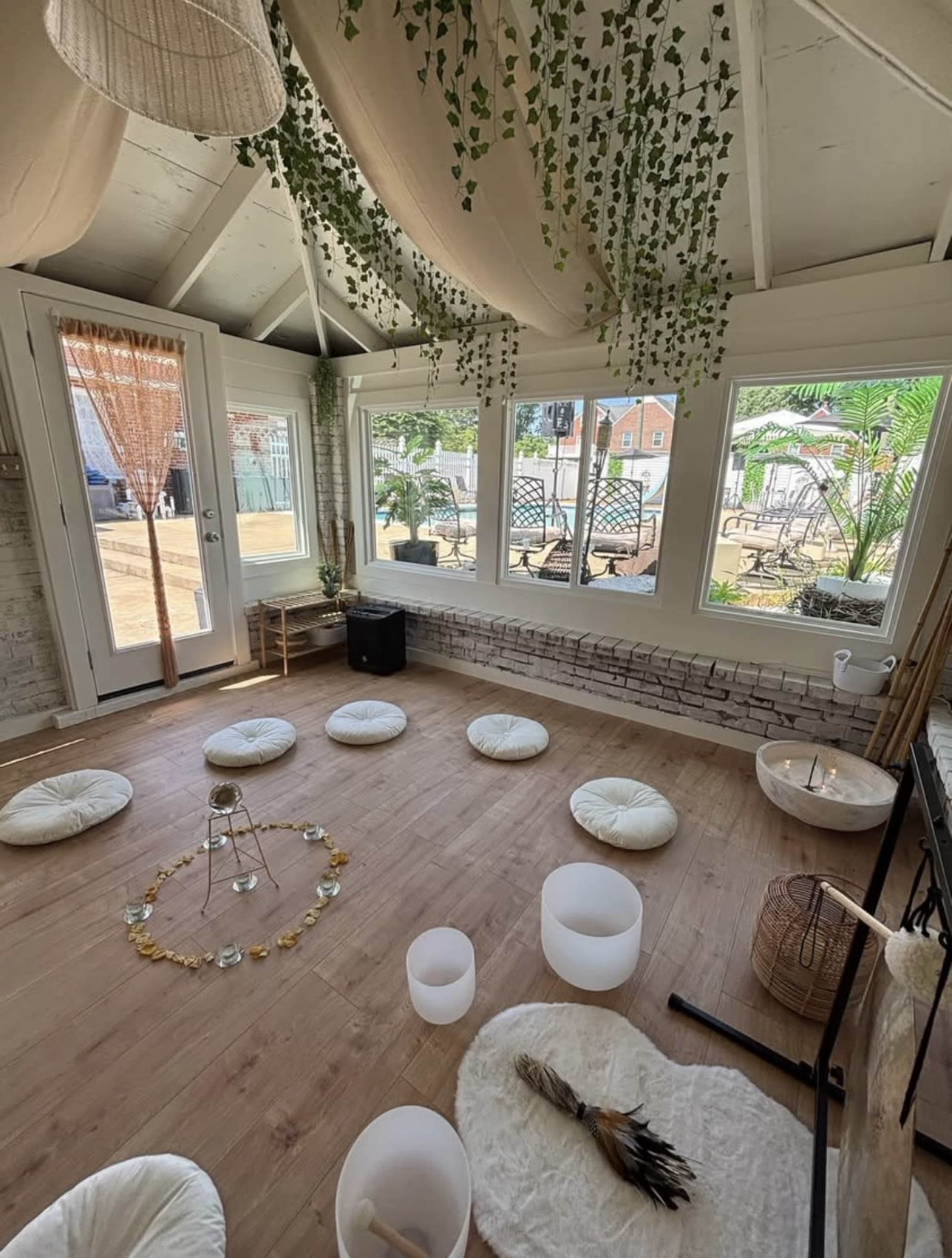 The image shows a serene indoor space with white cushions arranged in a circular pattern on a wooden floor, surrounded by large windows and greenery.