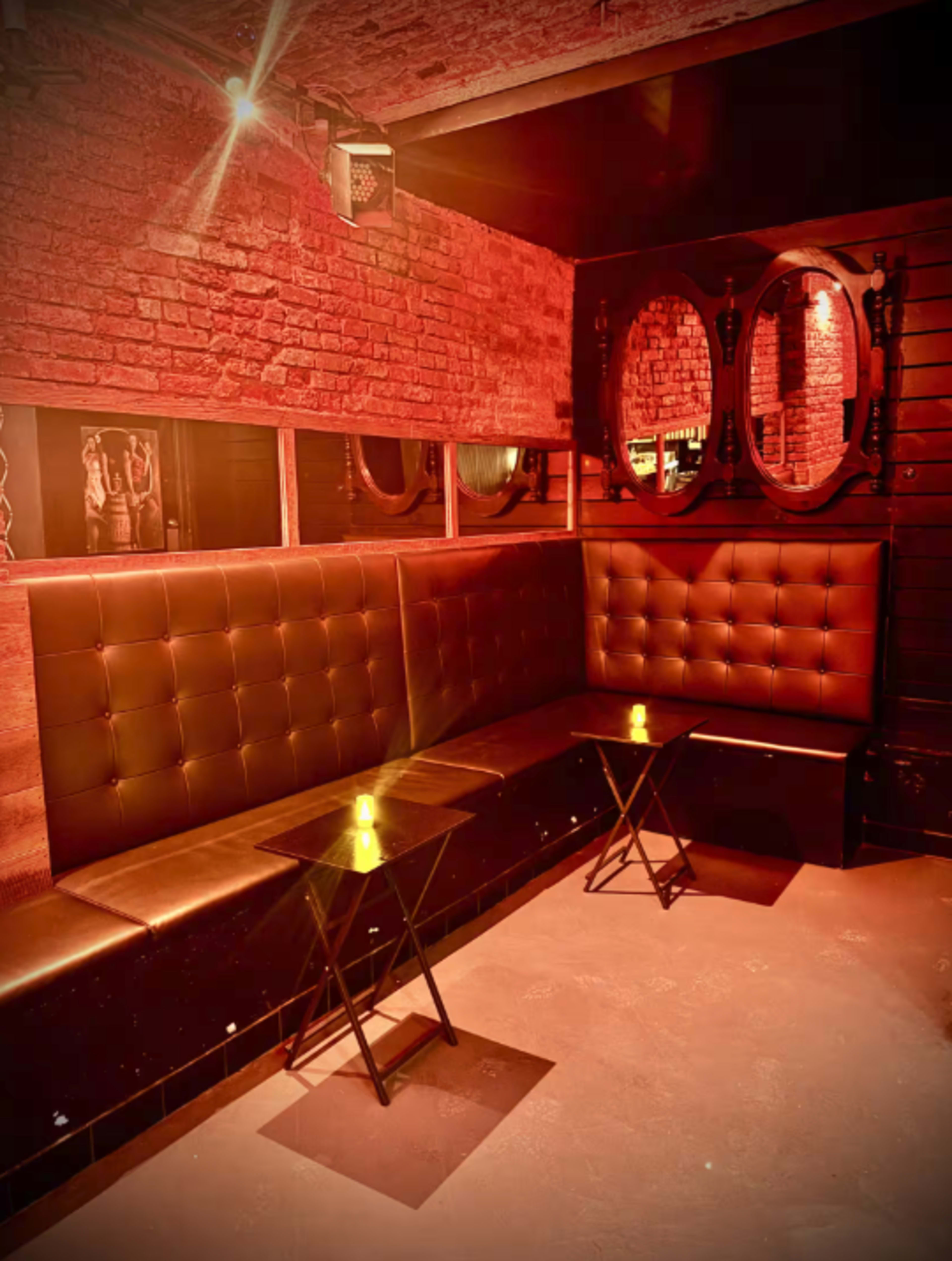 The image shows a dimly lit lounge area with red brick walls, leather seating, and small black tables near the seating.