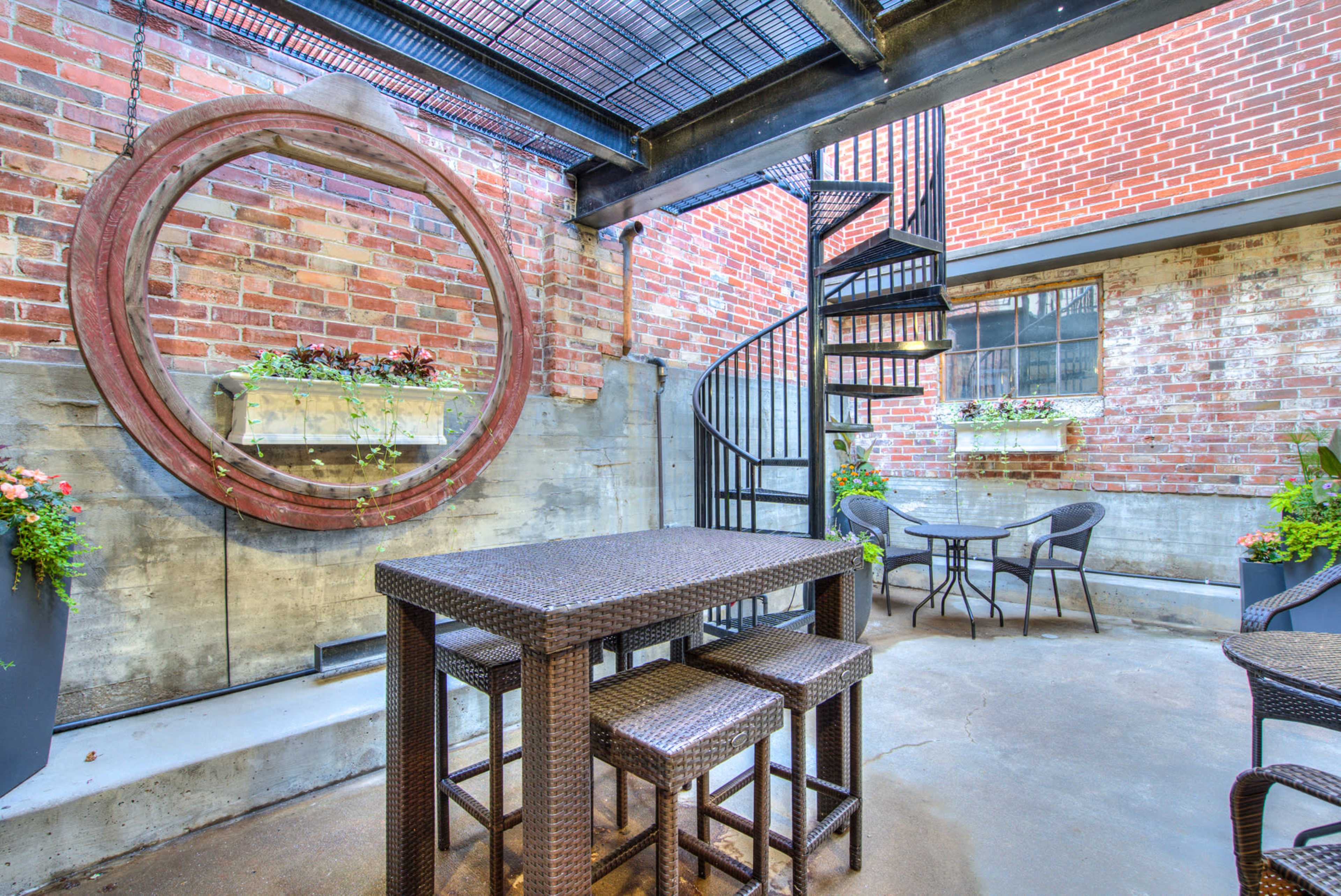A small outdoor patio with a spiral staircase, a dark wicker table and chairs, and brick walls adorned with planters.