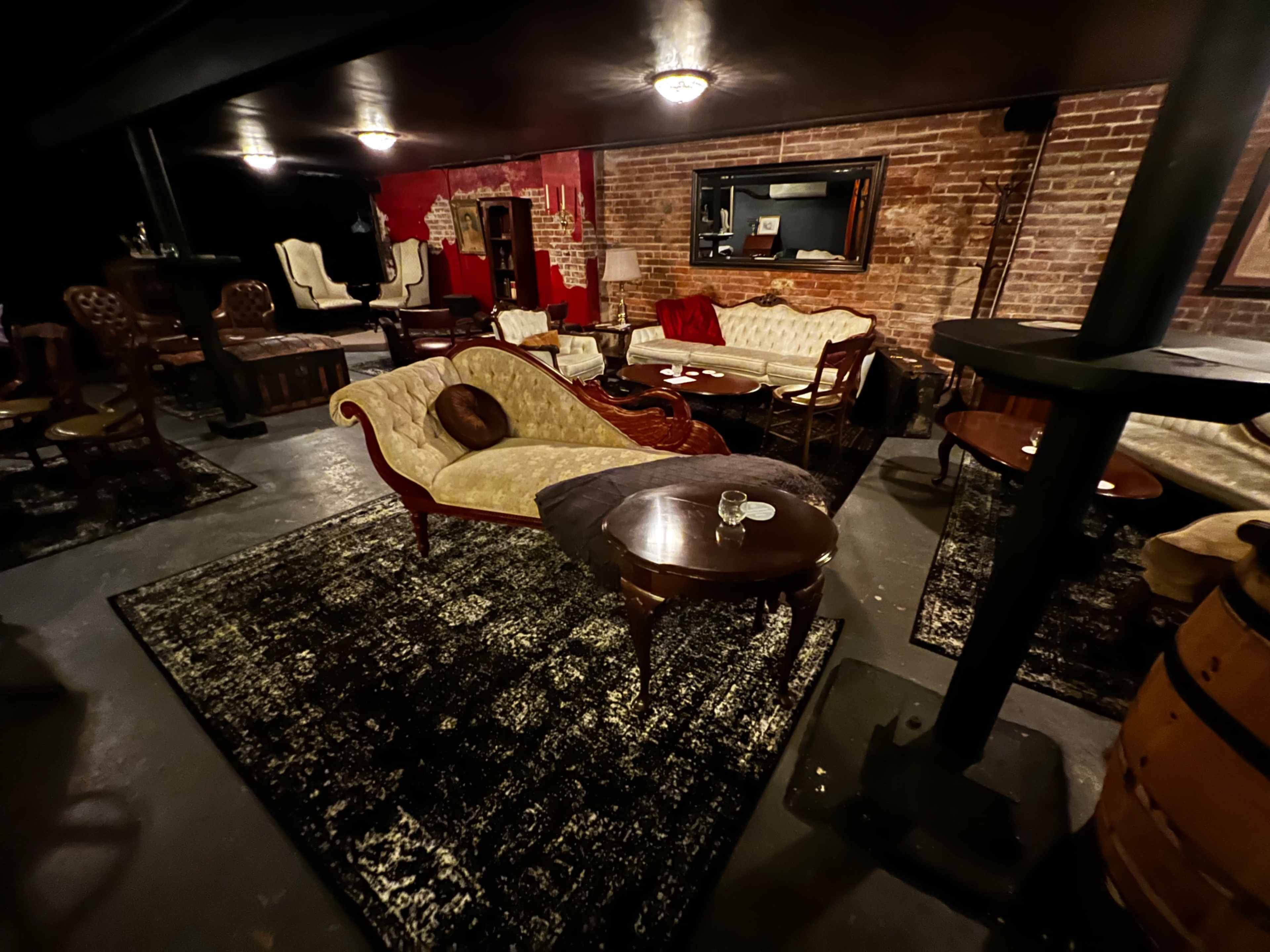 A dimly lit lounge area with vintage furniture, including a chaise longue, wooden tables, and exposed brick walls.