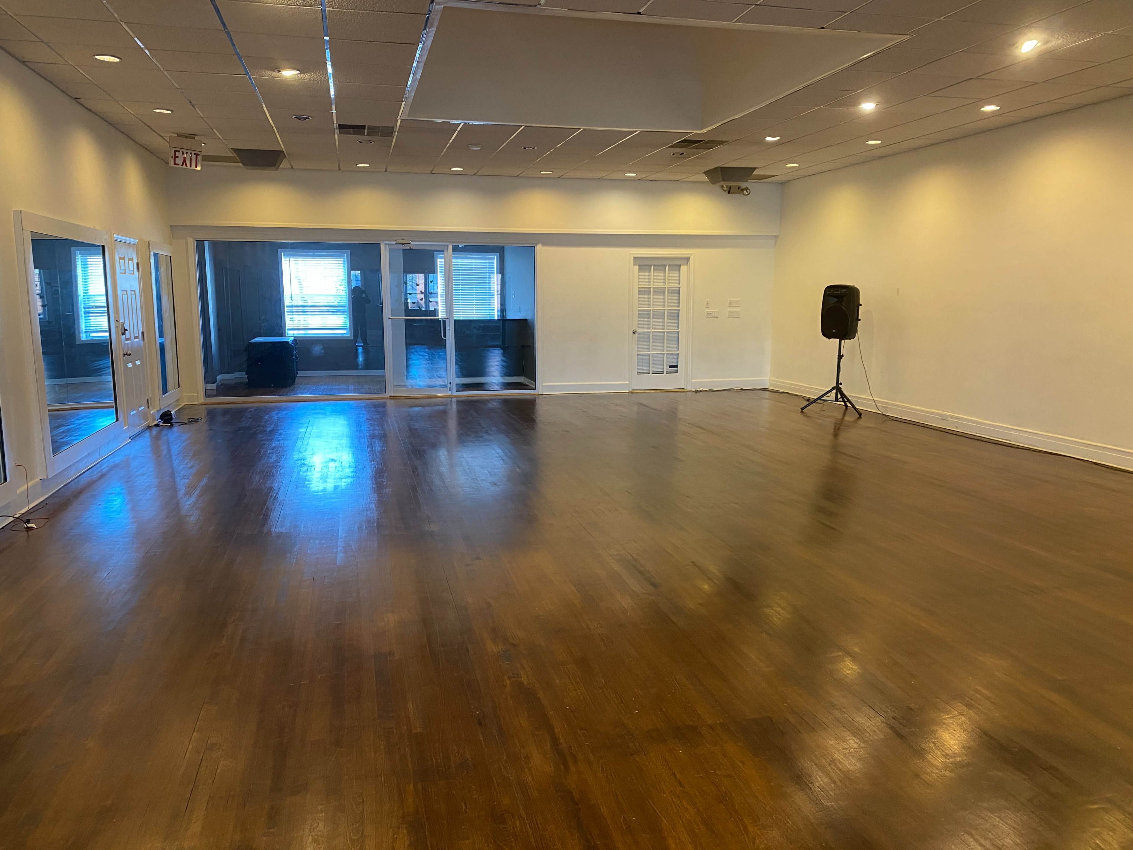 The image shows a large, empty room with hardwood floors and white walls, featuring two glass doors on one side and a speaker on a stand in the corner.