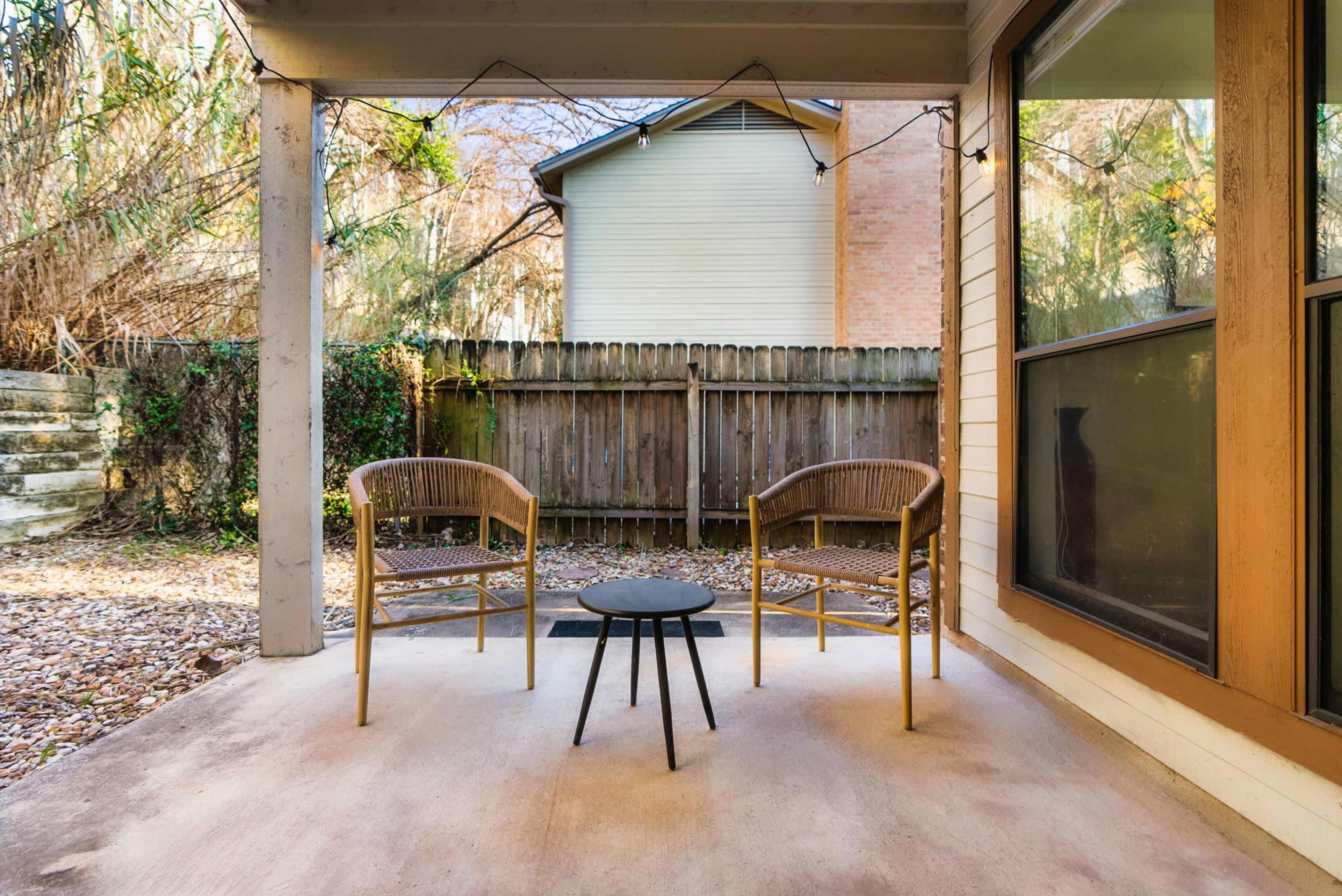 A covered patio features two chairs and a small table, surrounded by a backyard fenced with wooden planks.