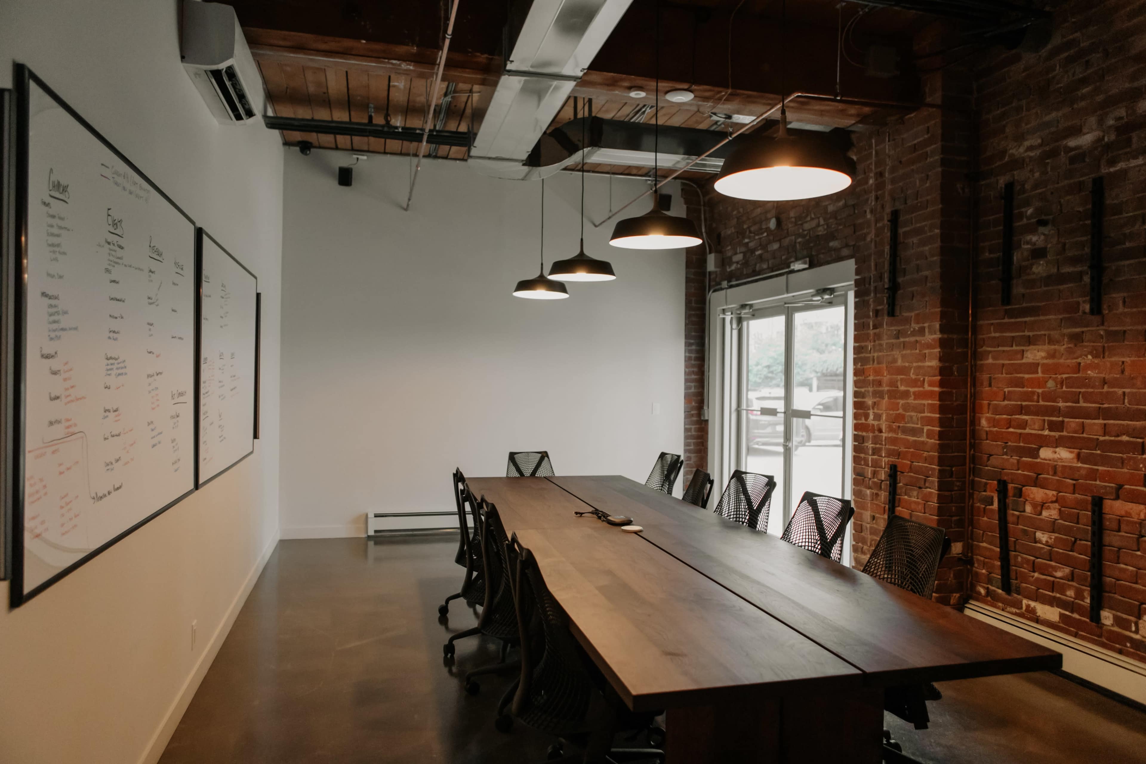 A large conference room features a wooden table surrounded by black mesh chairs, with whiteboards on the wall and industrial-style lighting overhead.