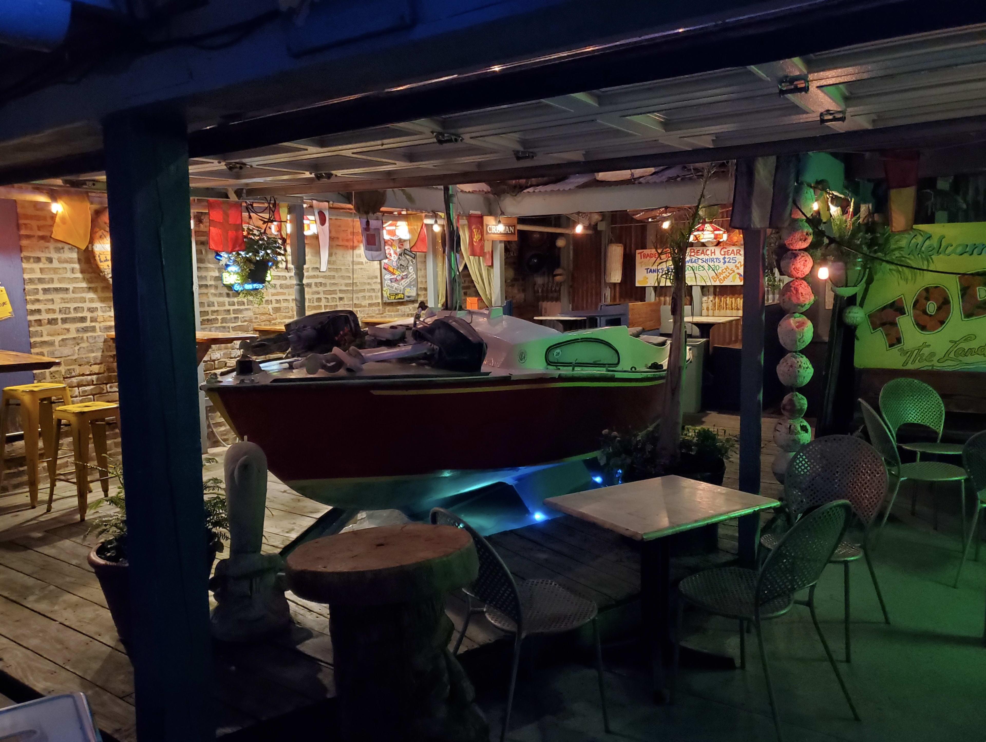 The scene features a colorful outdoor dining area with a boat displayed prominently in the center, surrounded by tables and chairs under soft lighting.
