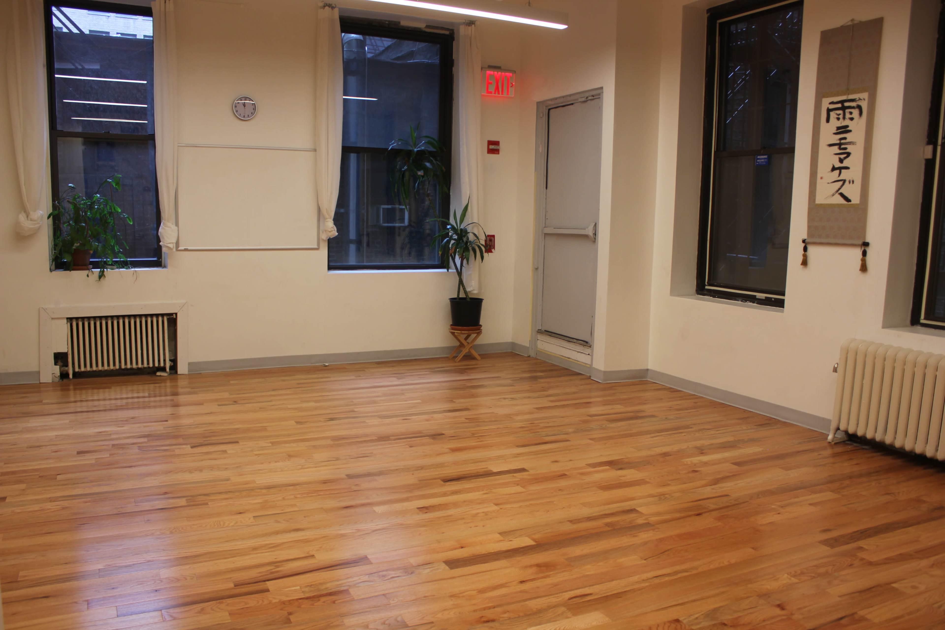 A spacious room with hardwood floors, large windows, a wall clock, and a potted plant.