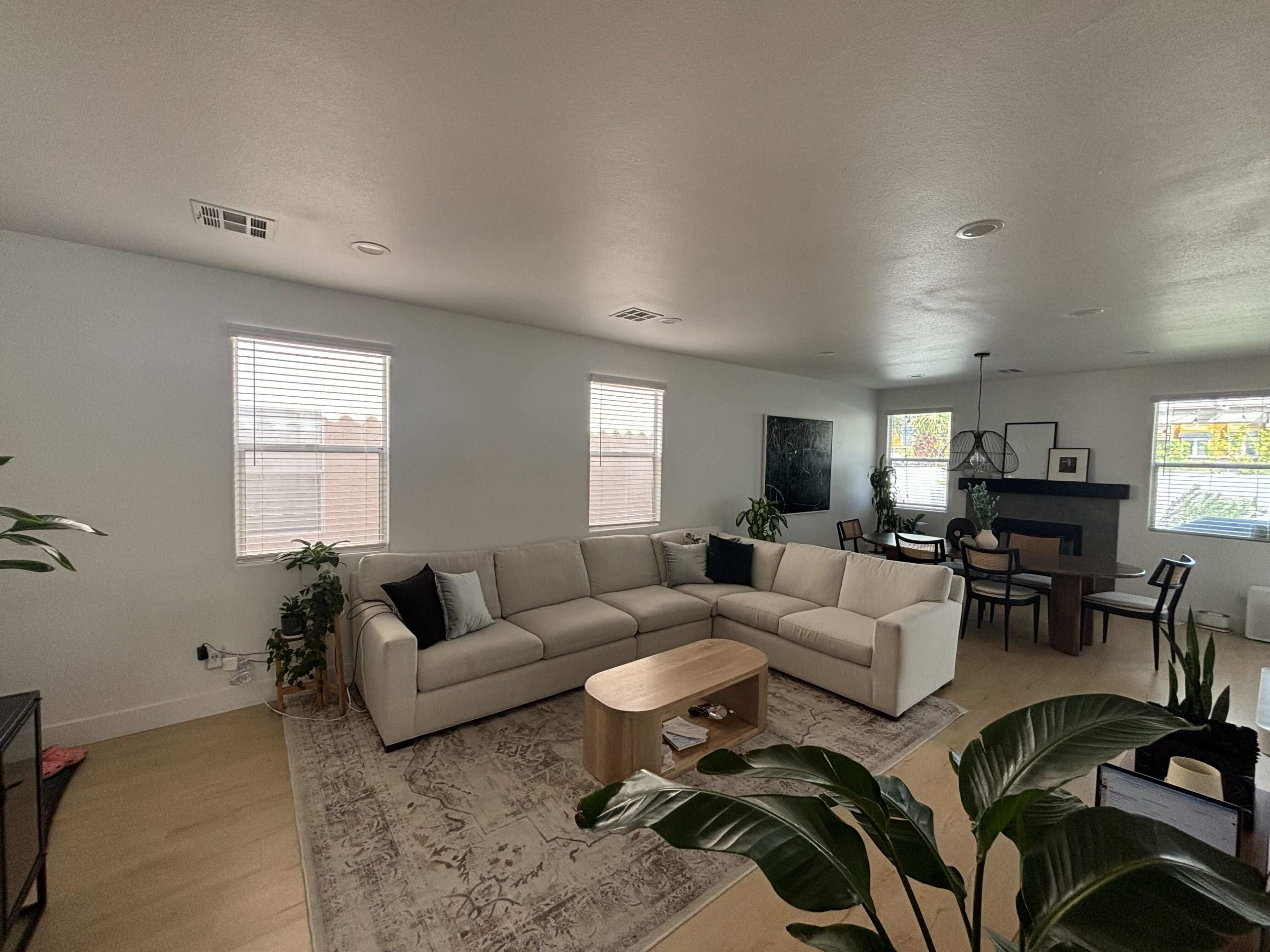 The image shows a modern living room with a sectional sofa, a coffee table, plants, and a dining area in the background.