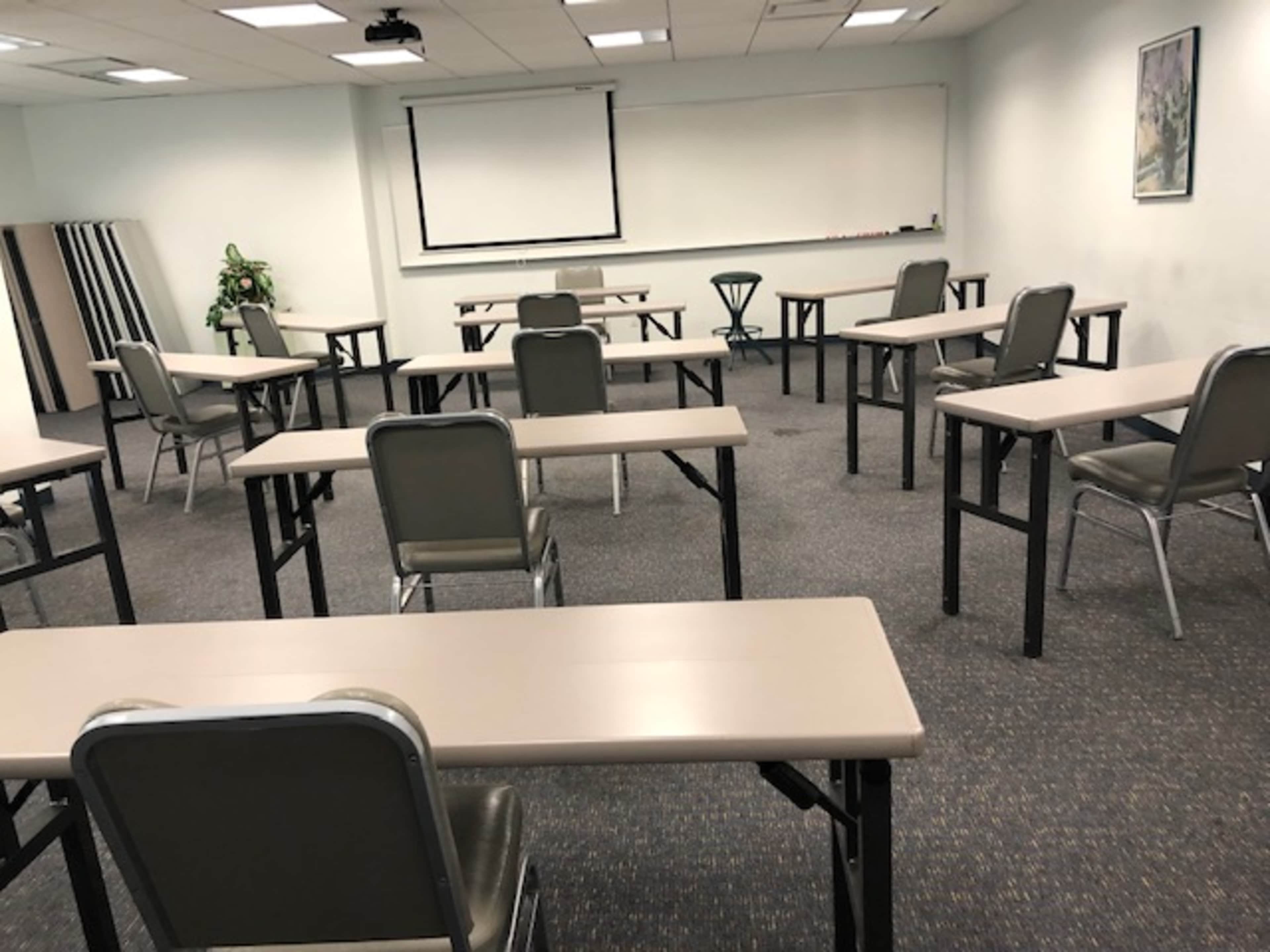 A classroom setup features several empty desks and chairs arranged in an open space with a projector screen at the front.