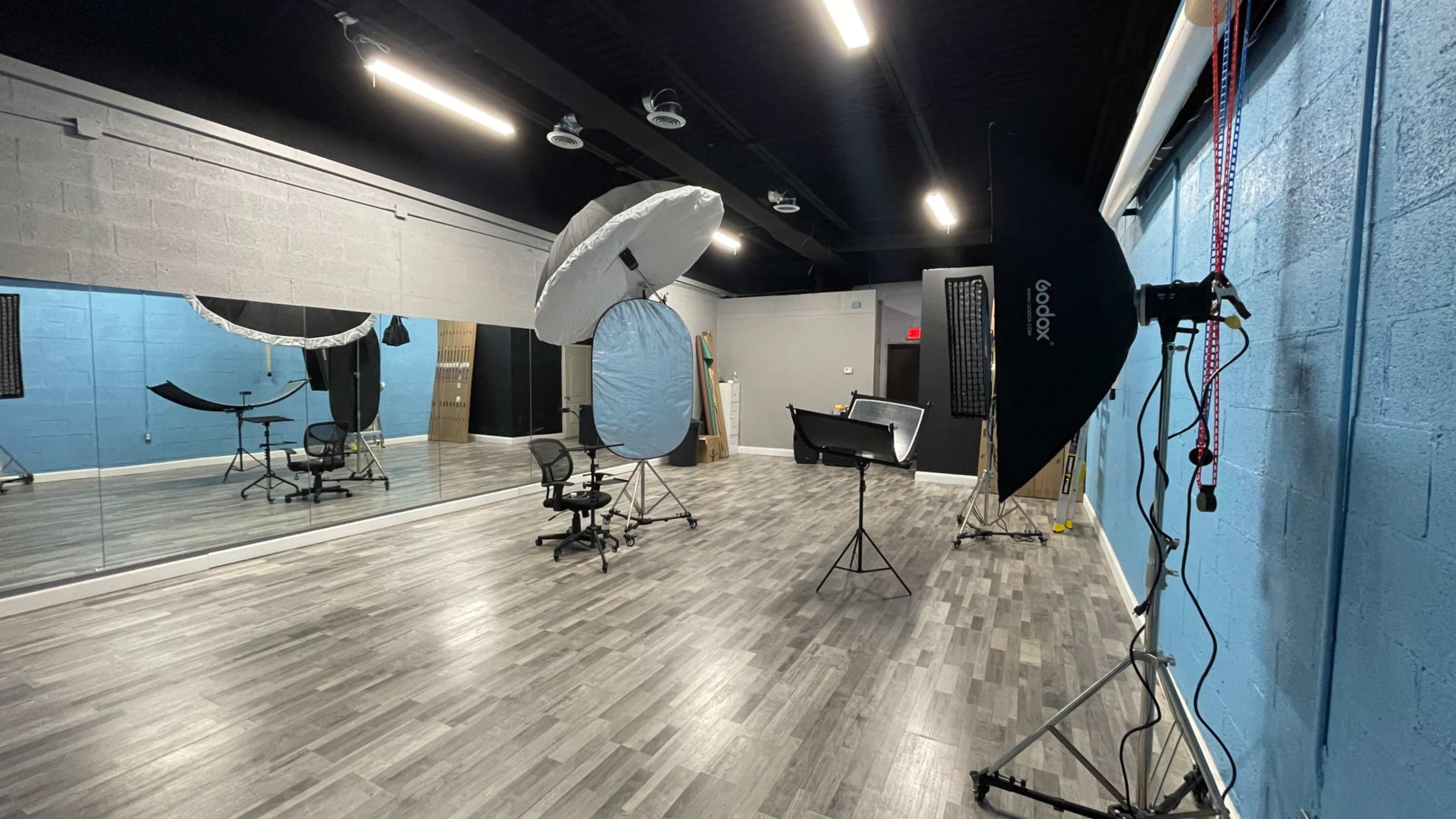 The image shows a photography studio with various lighting equipment, a mirrored wall, and a chair set up in a spacious room with blue and gray walls.