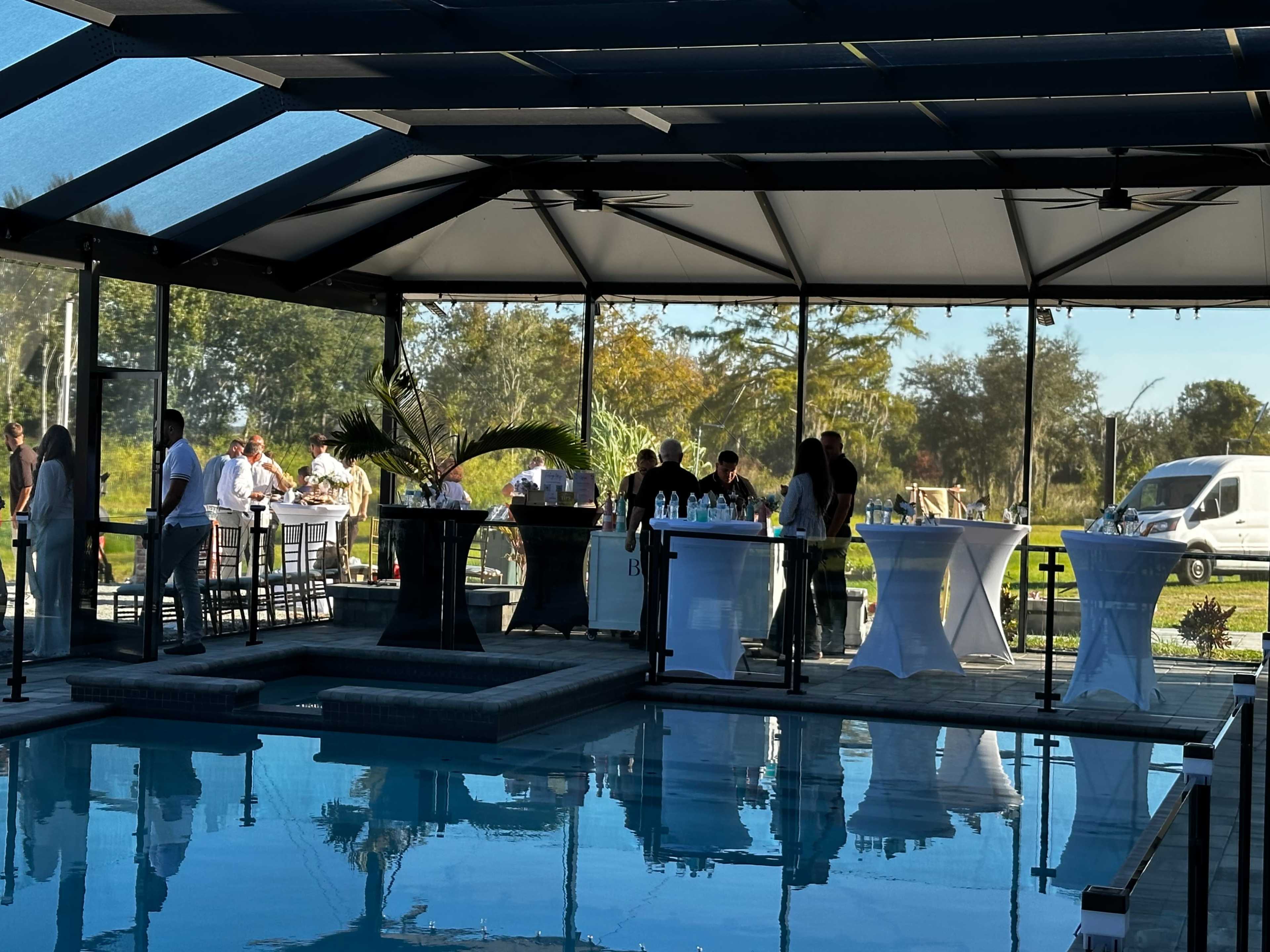 The scene shows a gathering of people inside a screened patio by a pool, with tables and a bar area set up for an event.