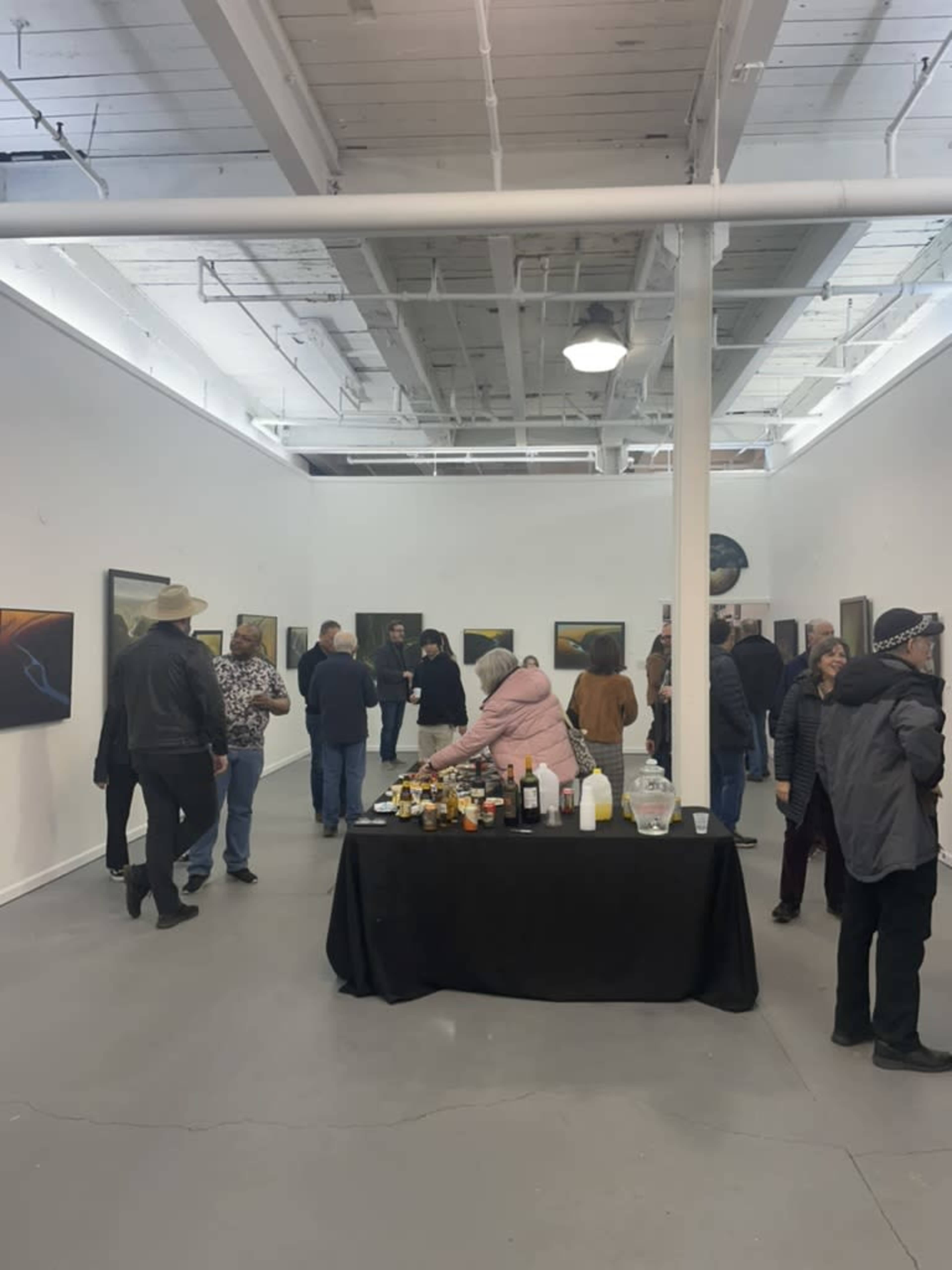 A group of people mingles in an art gallery featuring several paintings on white walls, with a table of refreshments in the foreground.