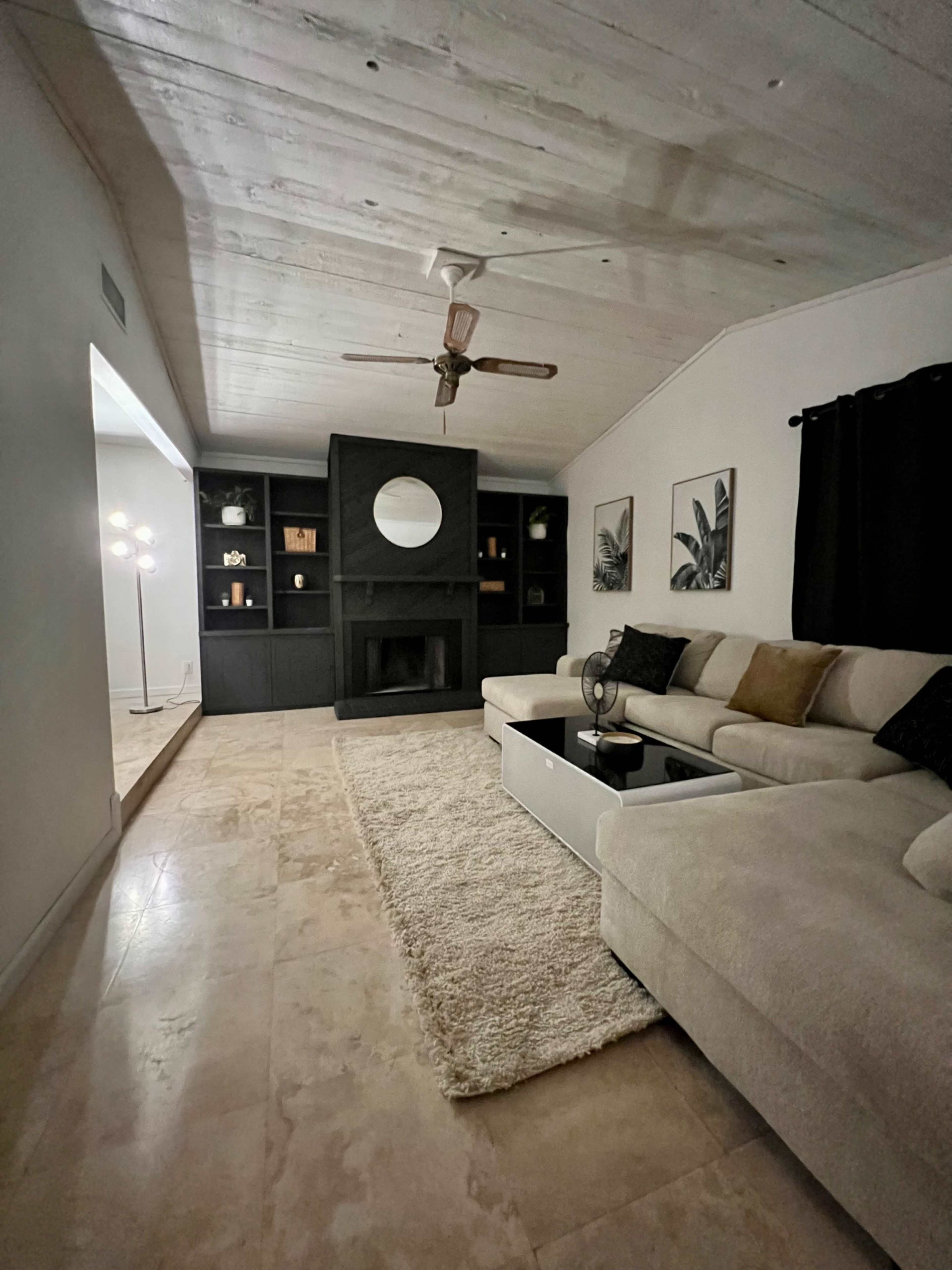 The image shows a living room with a light-colored sectional sofa, a coffee table, a dark feature wall with a circular mirror, and shelves filled with decor.